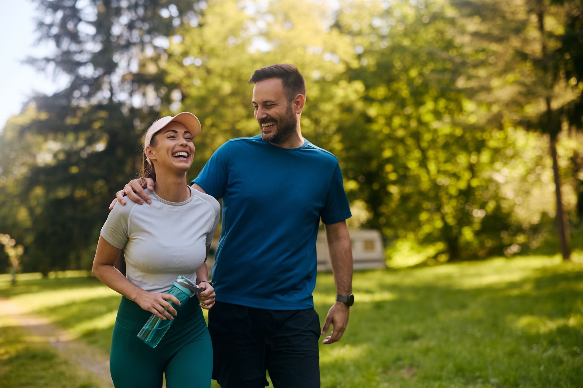 Two people walking and smiling in a sunny park, one holding a water bottle