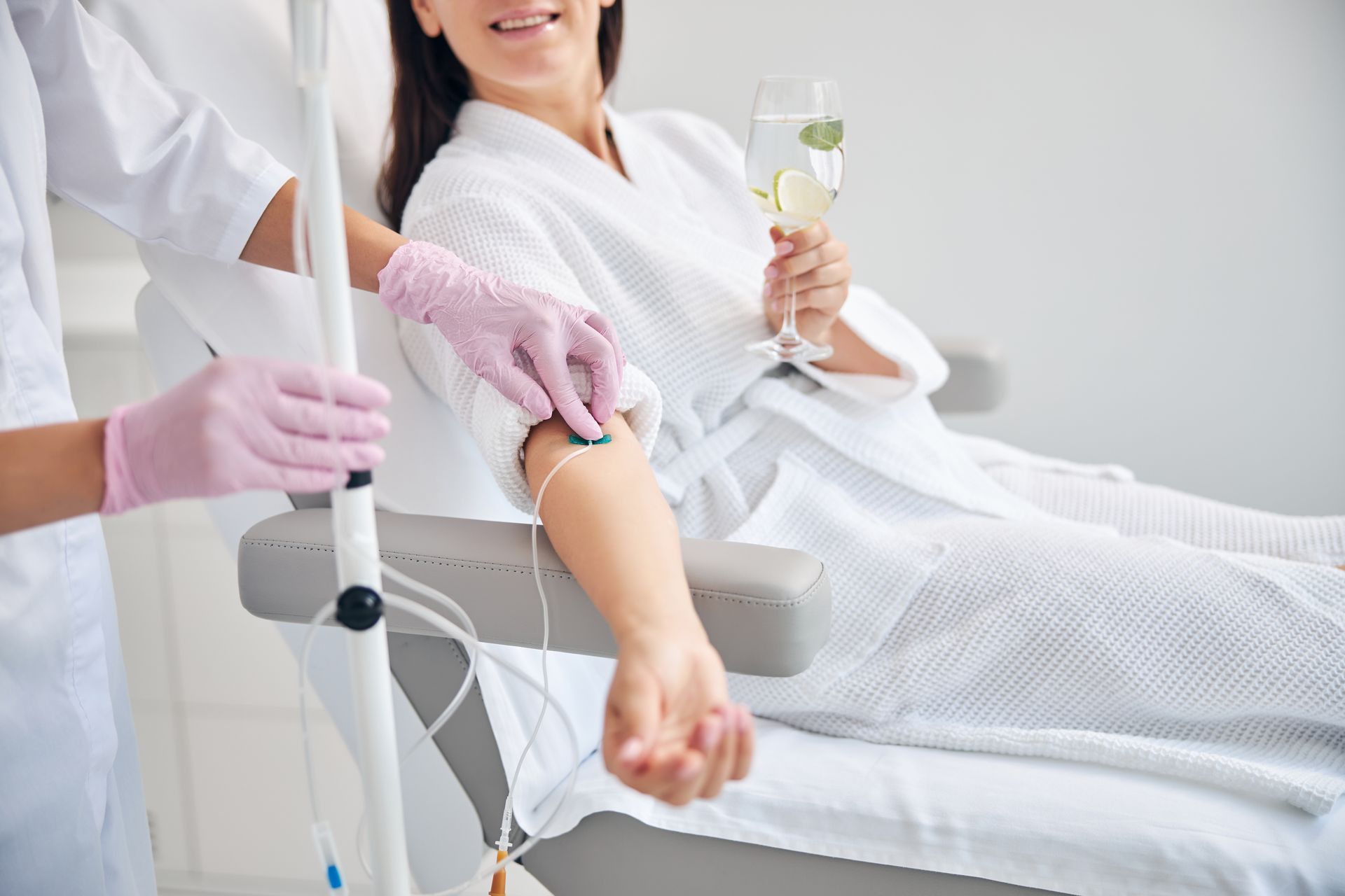 Woman in white robe receives IV drip in medical setting, holding drink and smiling.