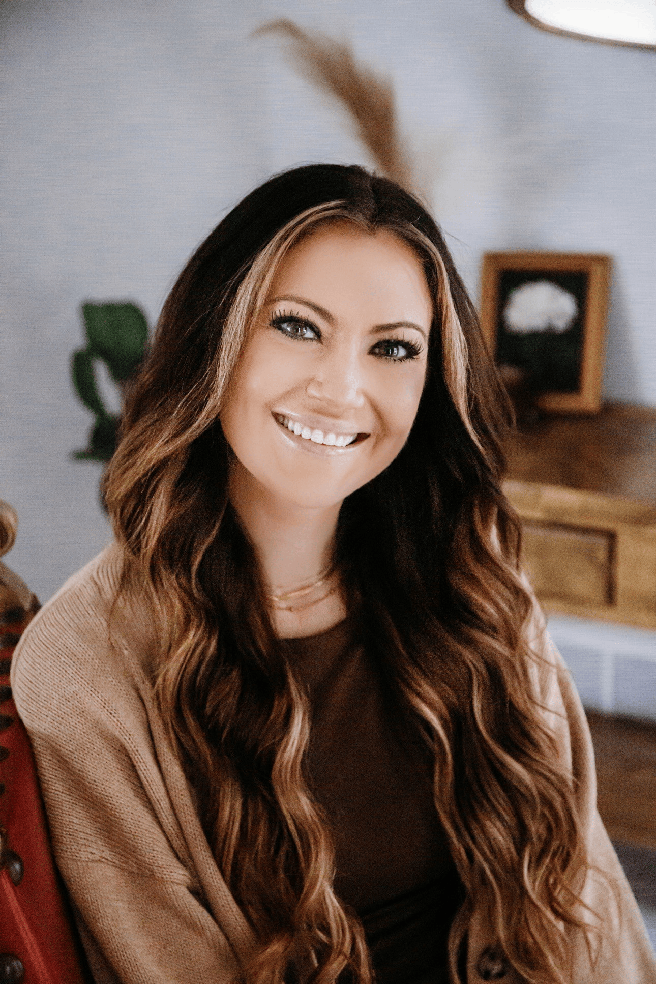 Smiling woman with long wavy brown hair in a cozy indoor room, wearing a brown sweater.
