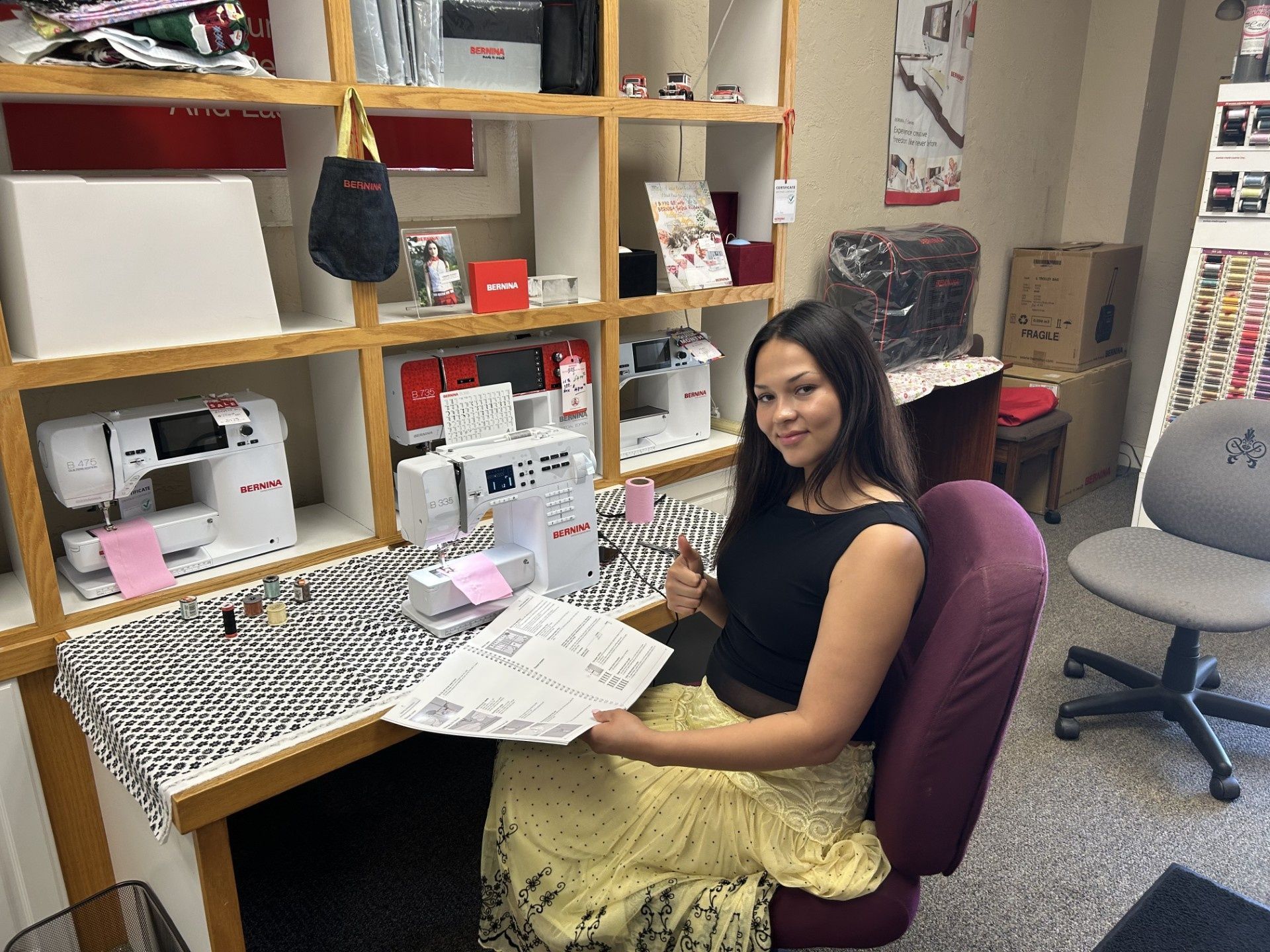 Woman seated at a desk in a craft room, holding papers beside organized shelves and sewing supplies