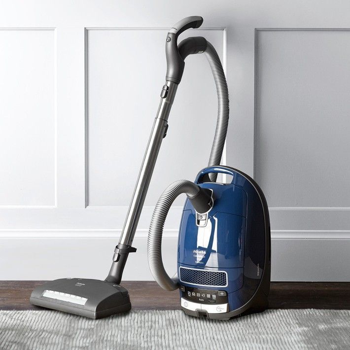 Blue canister vacuum cleaner with hose and floor brush on a gray carpet against a white wall