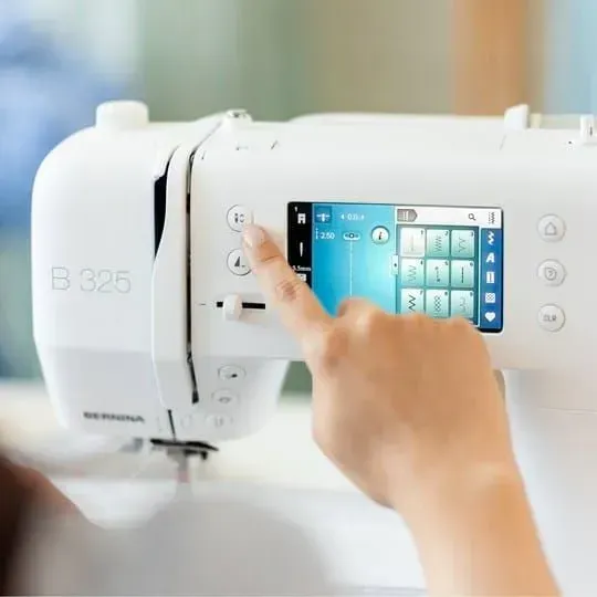 Hand using a smartphone to control a white sewing machine with touchscreen controls