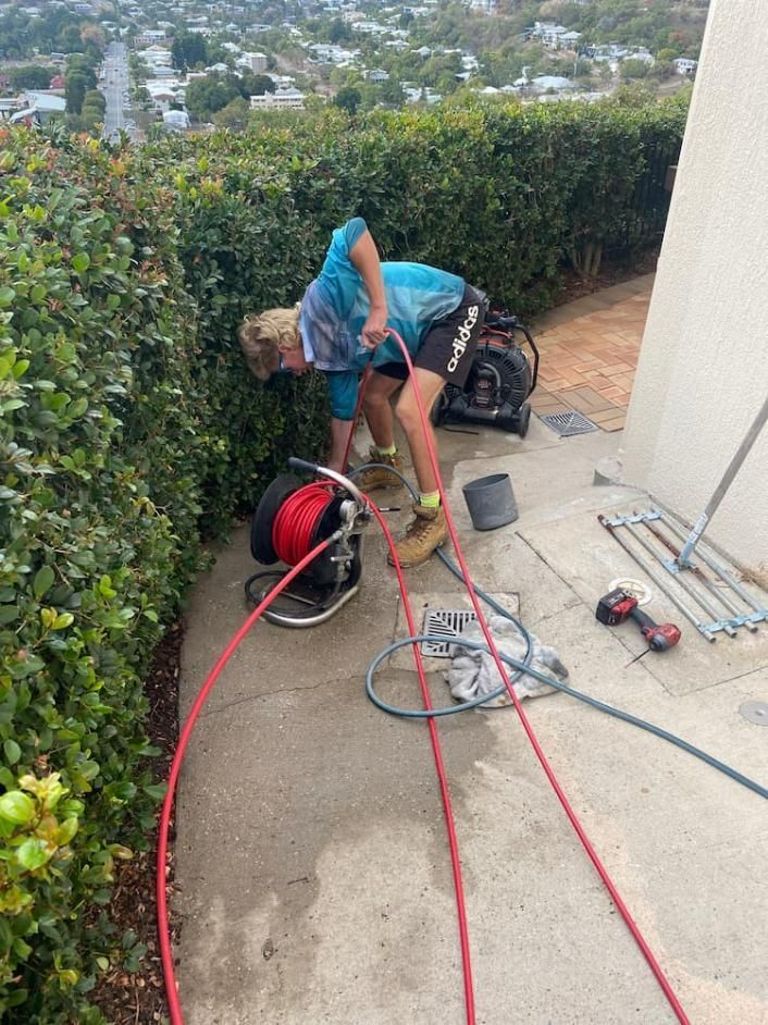 Man clearing a drain with red hose. Outdoor setting with green foliage and city view — Top To Bottom Plumbing in Garbutt, QLD