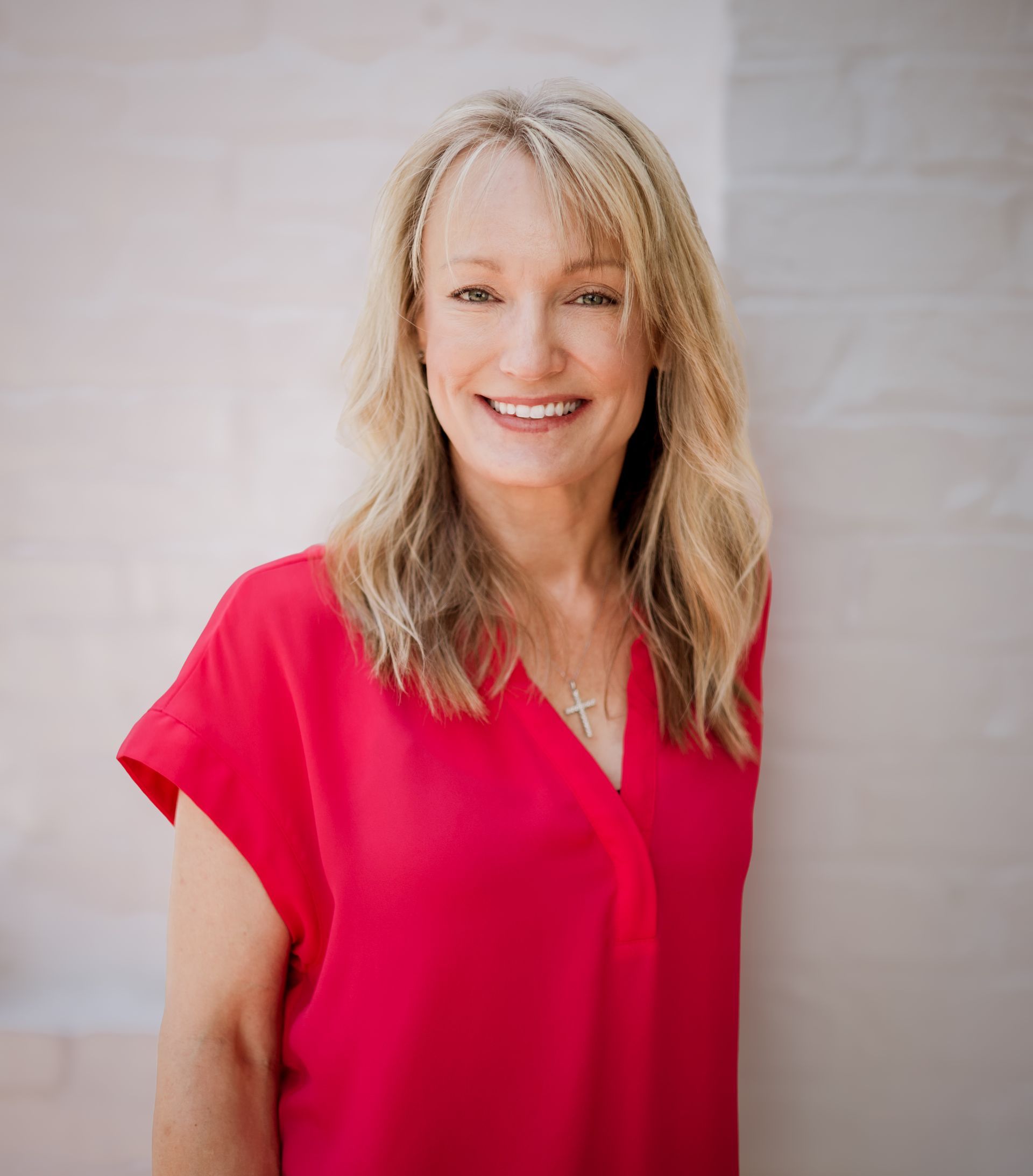 A smiling woman with blonde hair, wearing a vibrant red top and a cross necklace, stands against a white brick wall.