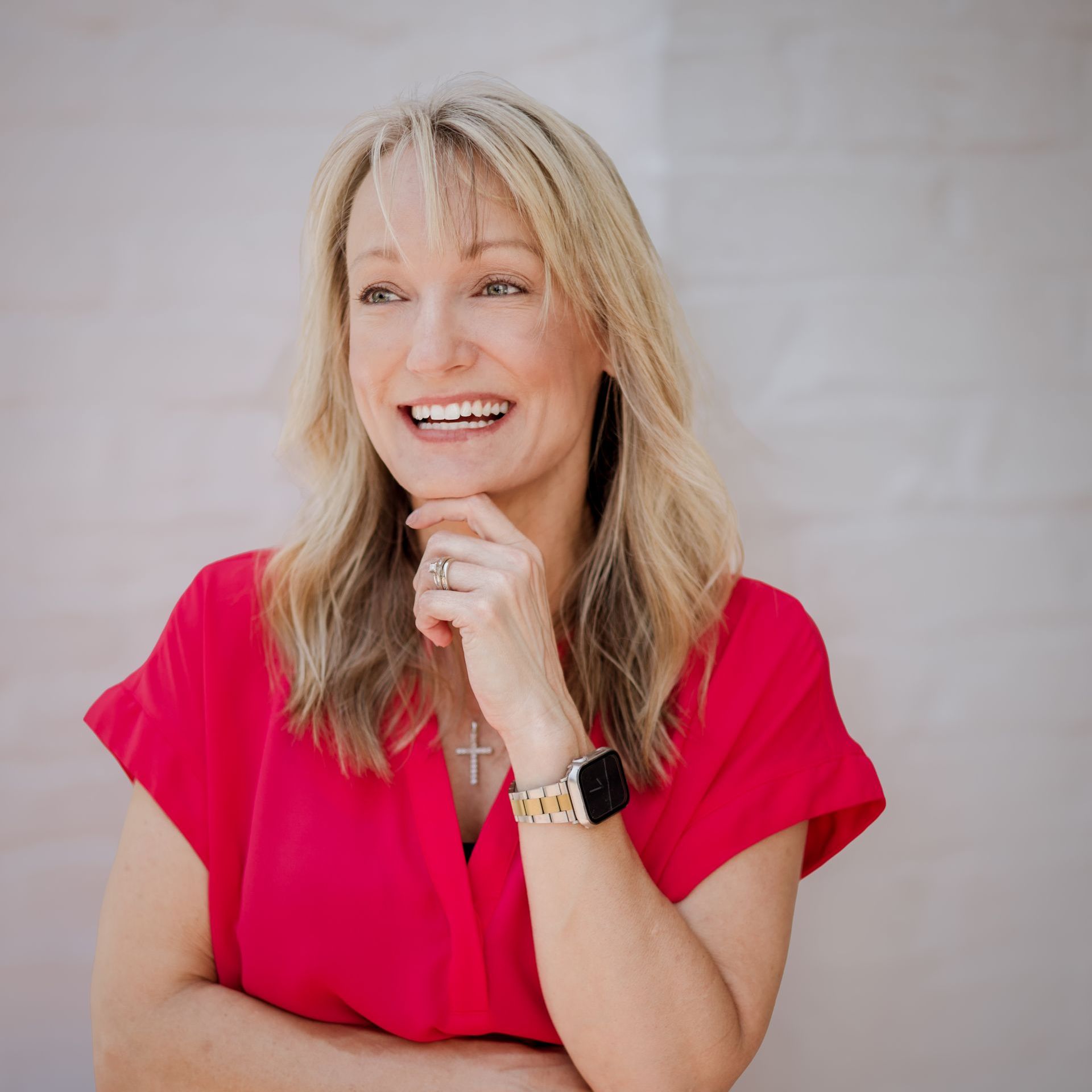A woman with blonde hair smiling while wearing a bright pink shirt, standing in front of a light-colored brick wall.
