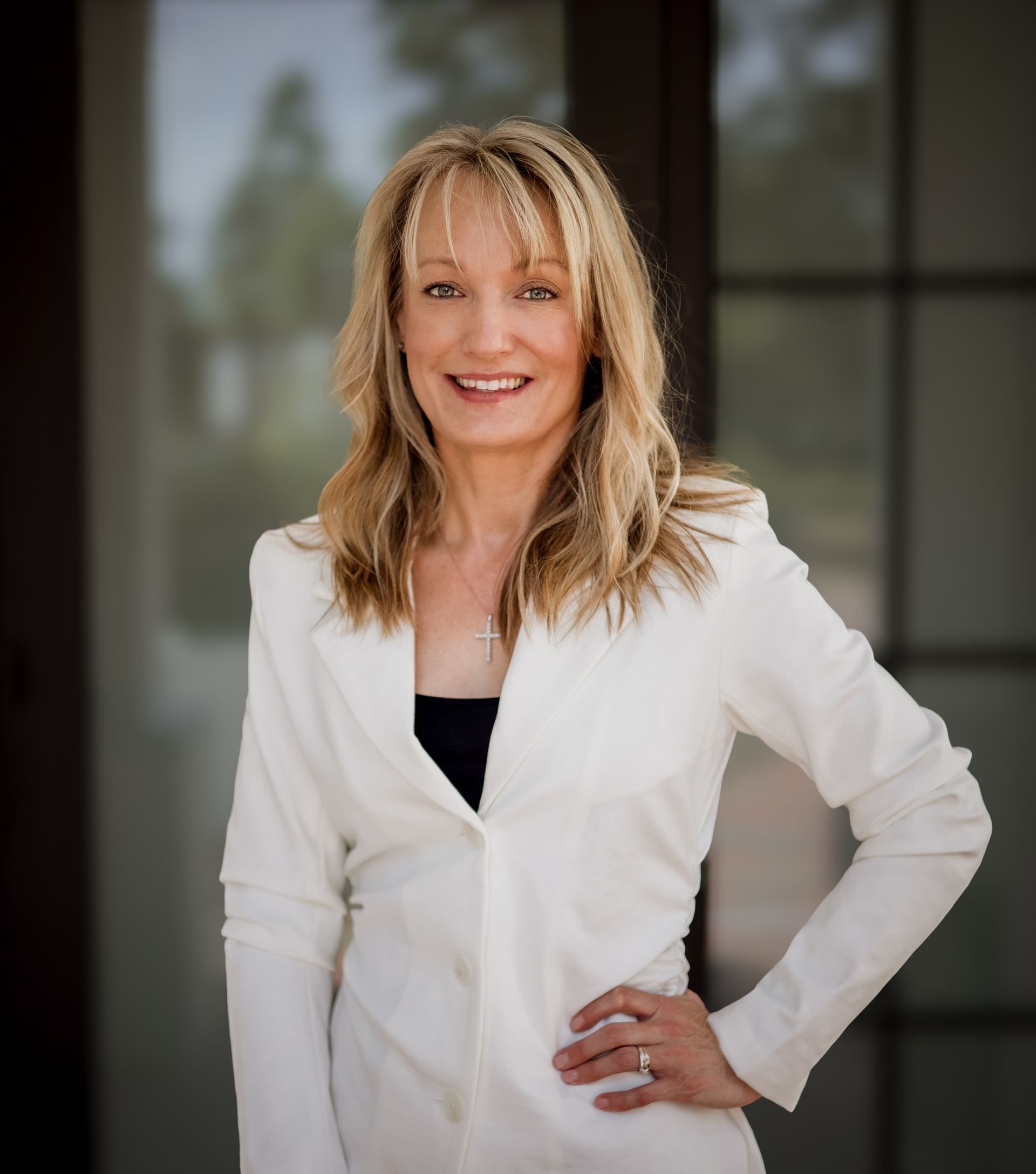A blonde person with a friendly smile, wearing a white blazer over a black top, poses with a hand on their hip.