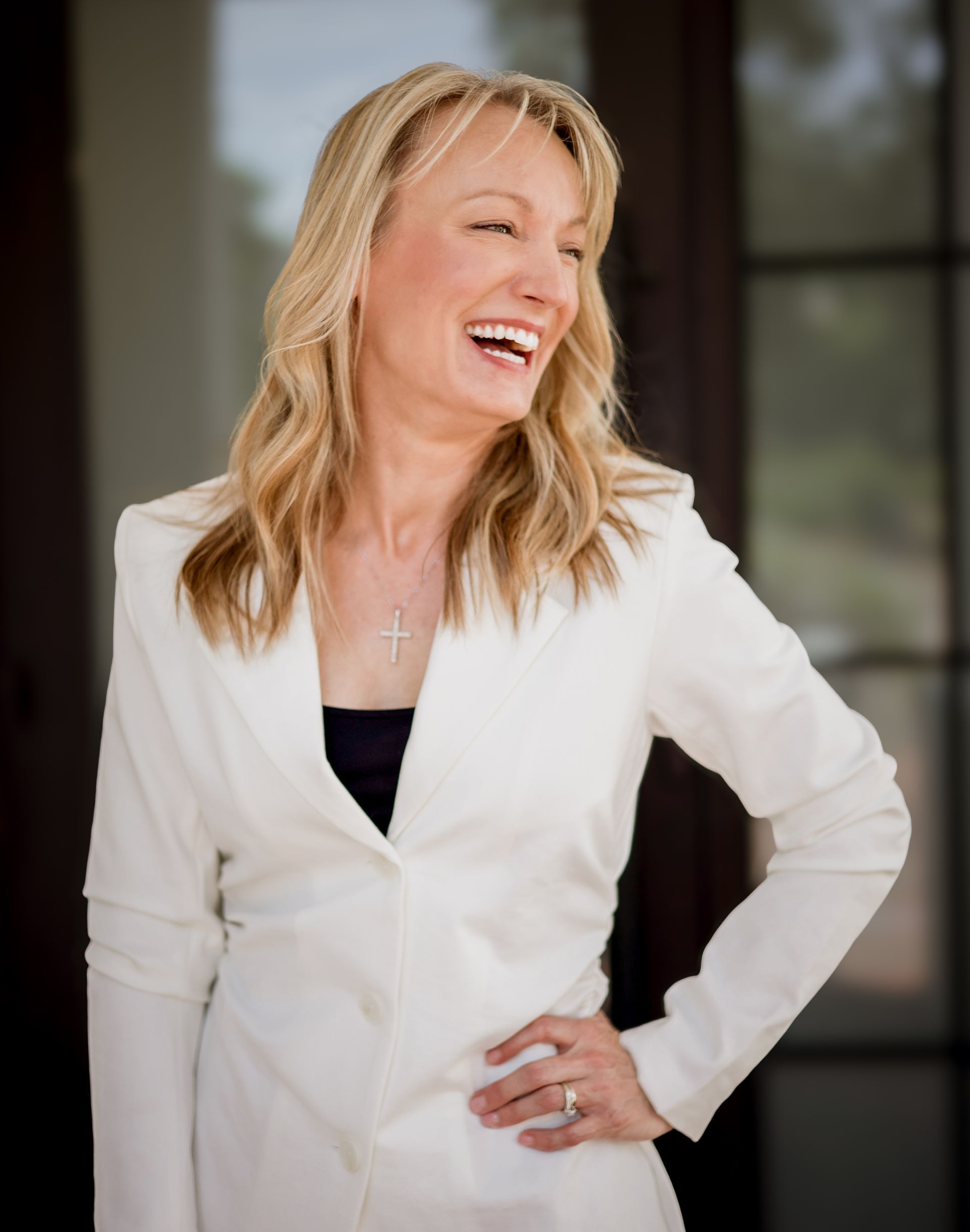 A person with blonde hair wearing a white blazer smiles warmly with one hand on their hip.