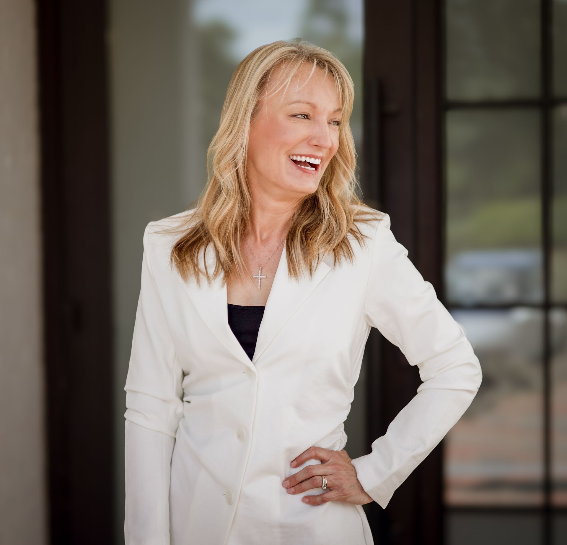 A smiling person with shoulder-length blonde hair, wearing a white blazer and a dark top, poses with a hand on their hip.
