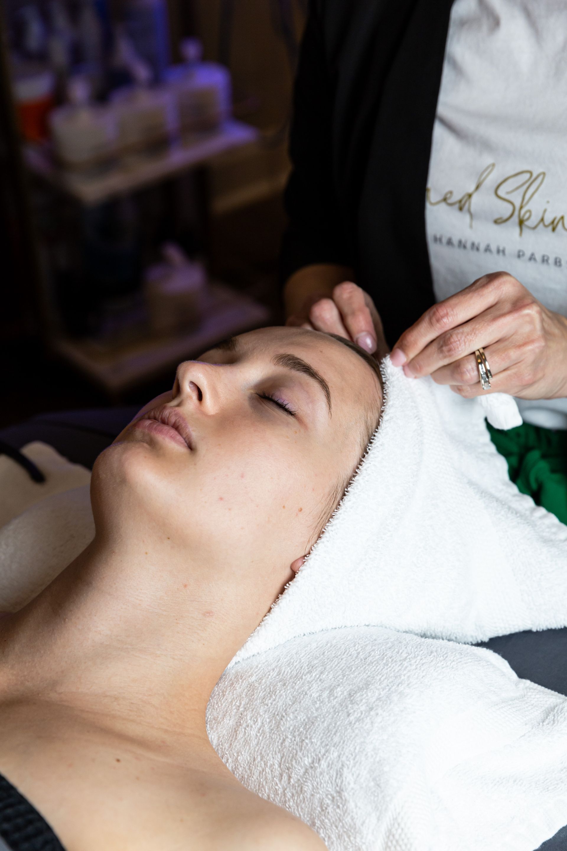 A person receiving a facial treatment with a white towel wrapped around their head, attended by an esthetician.
