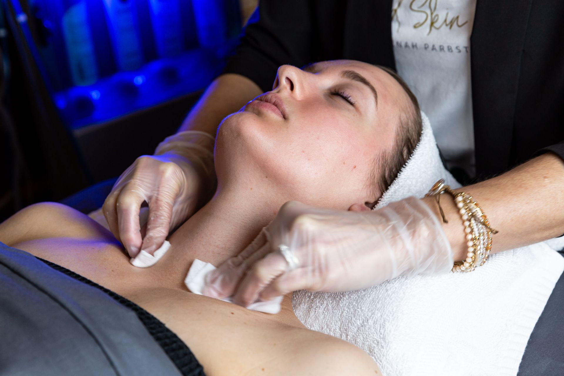 A professional esthetician wearing gloves cleanses a client's neck and chest area with cotton pads in a spa setting.