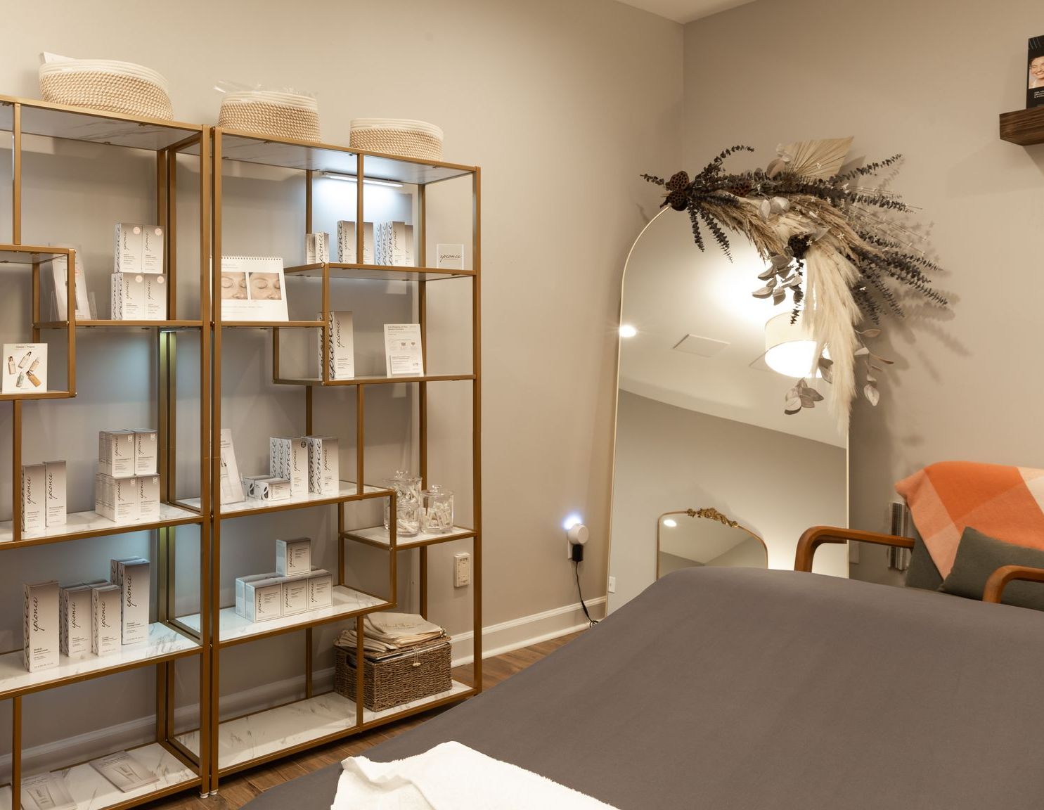A spa room featuring a gold metal shelving unit stocked with skincare products next to a large arched mirror with decor.