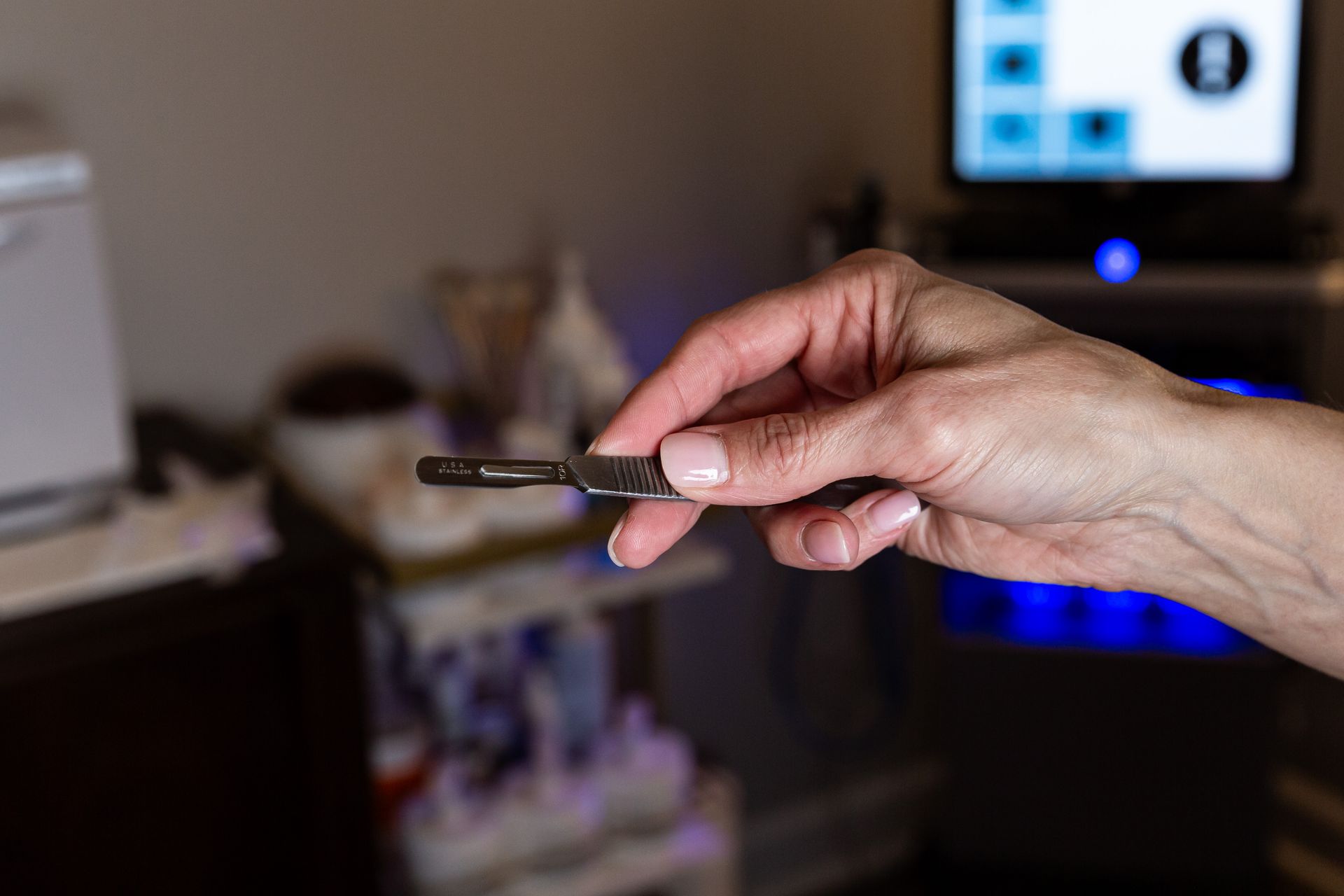 A hand holds a surgical scalpel in a medical office setting with equipment in the background.