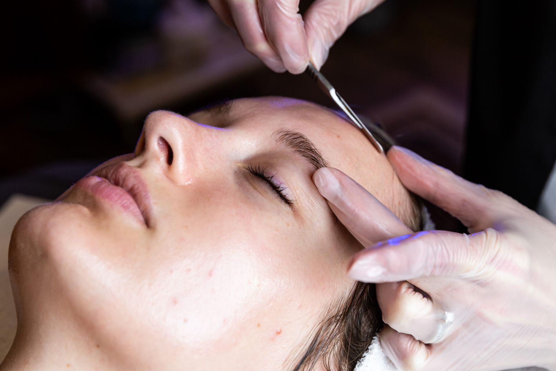 A person having their eyebrows shaped with a metal tool by a professional wearing gloves.