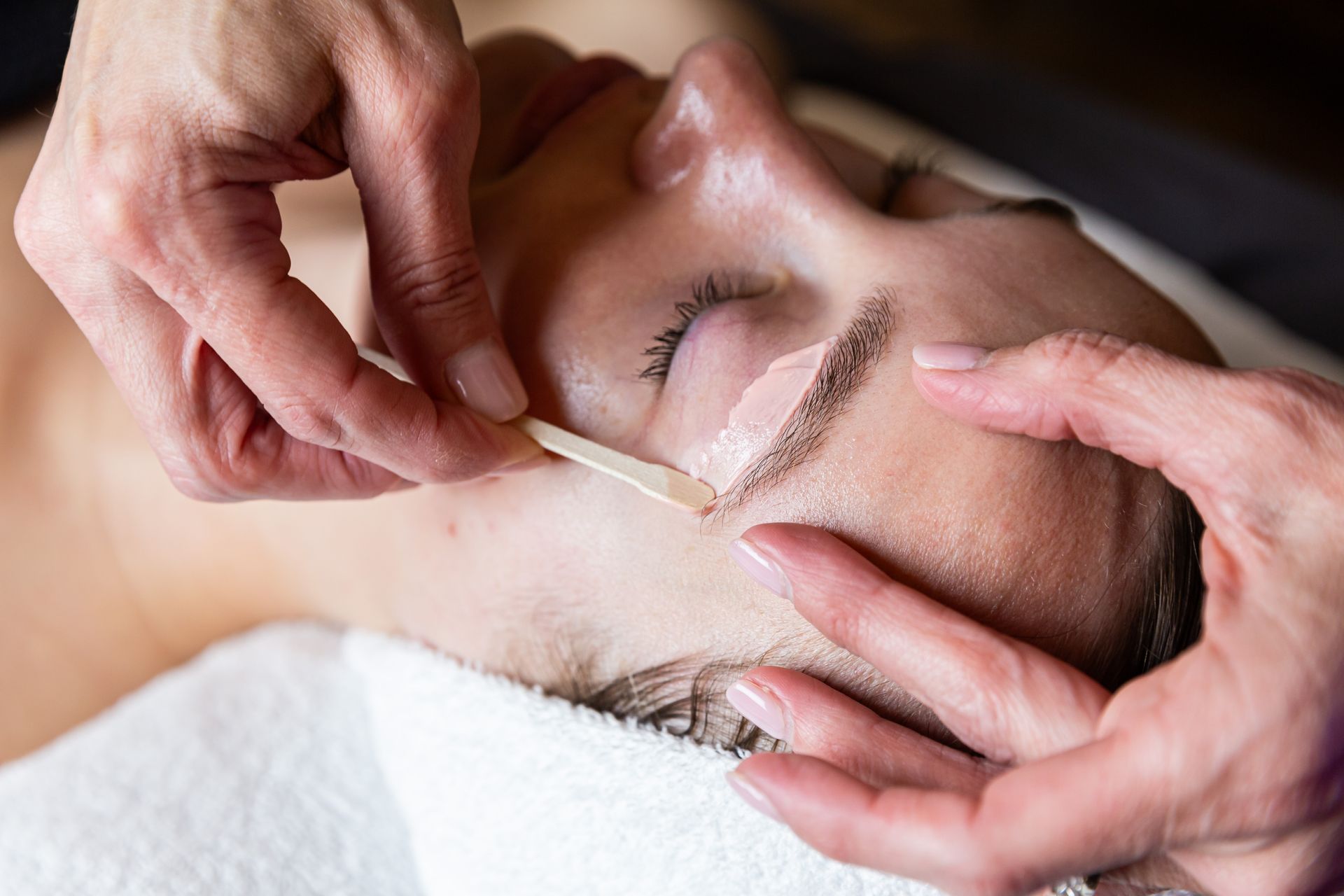 A beautician uses a wooden applicator to apply wax to a client's eyebrow in a salon setting.