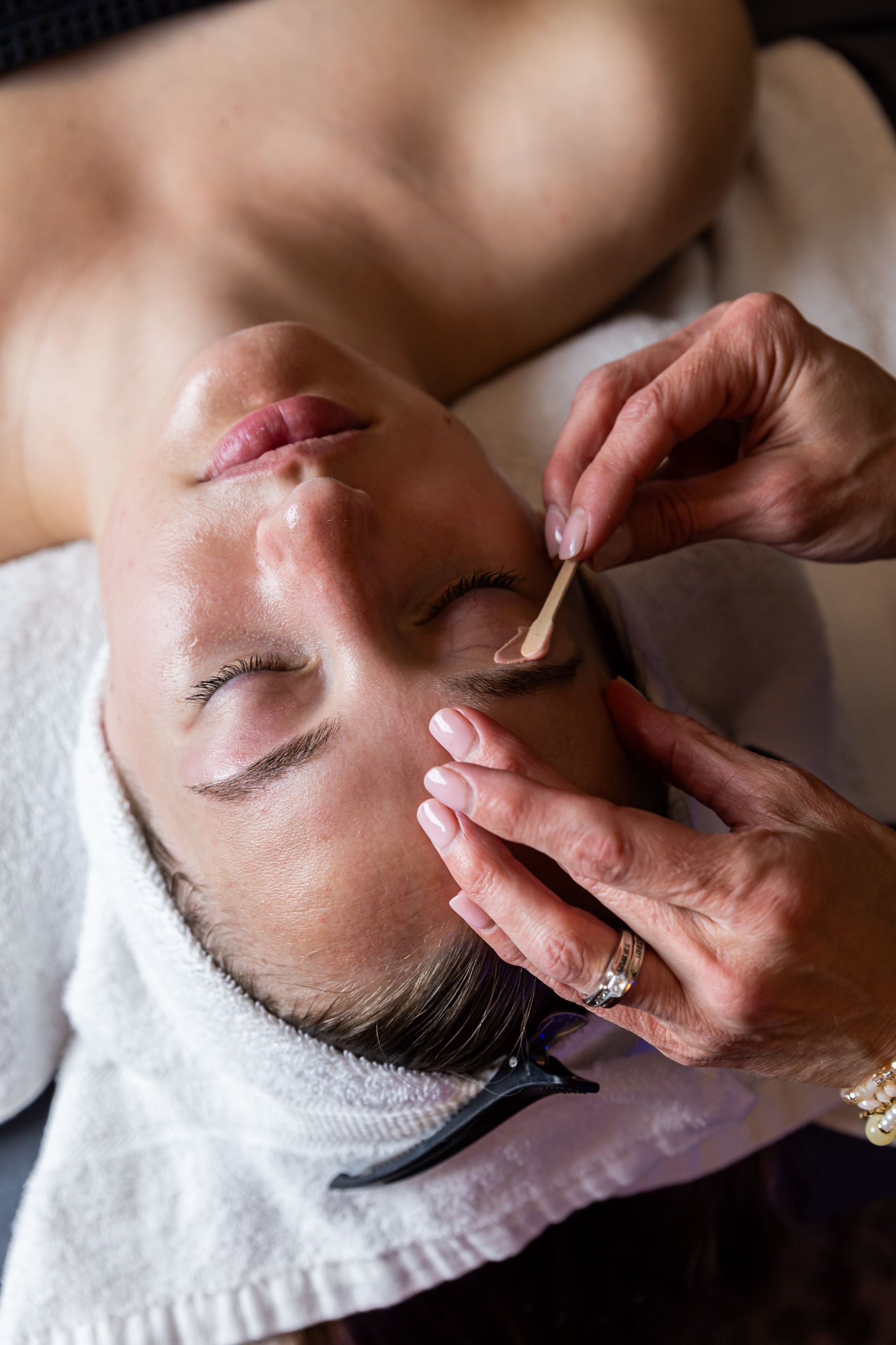 A person receiving a brow wax treatment from a professional with an applicator stick while lying on a salon table.