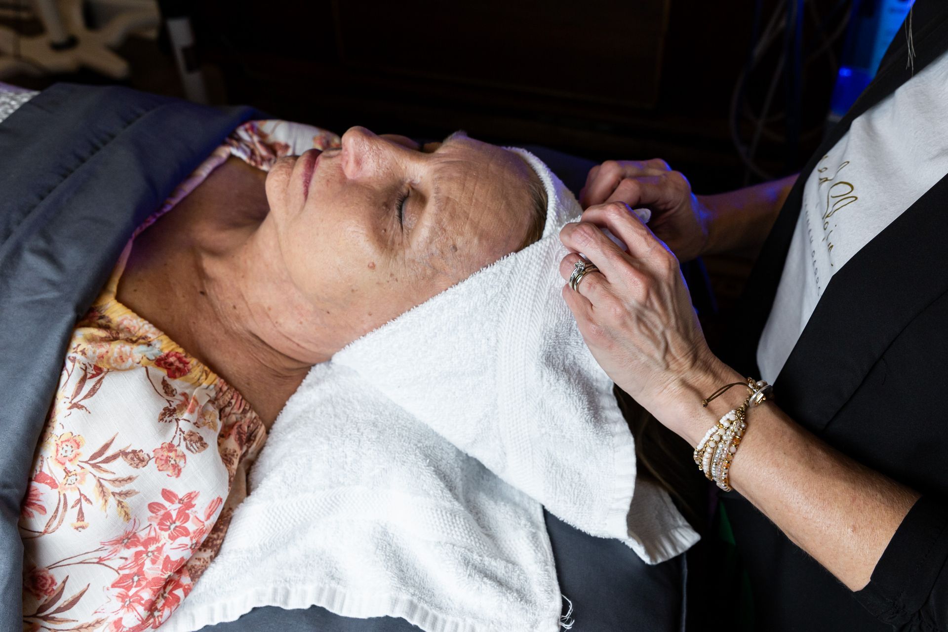 A person lies on a spa table while an aesthetician adjusts a white towel headband over their forehead.