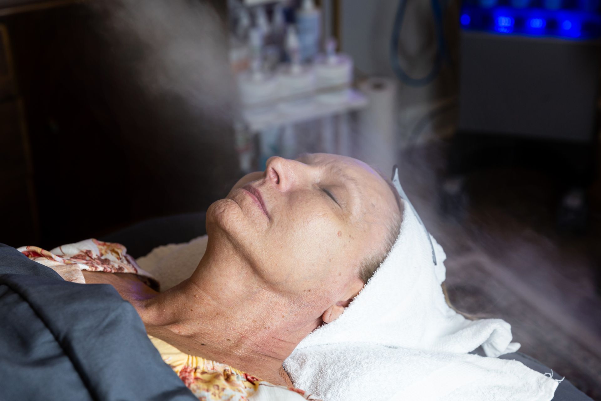 A person reclines with eyes closed under a facial steamer in a spa setting, wearing a white headband and gray drape.