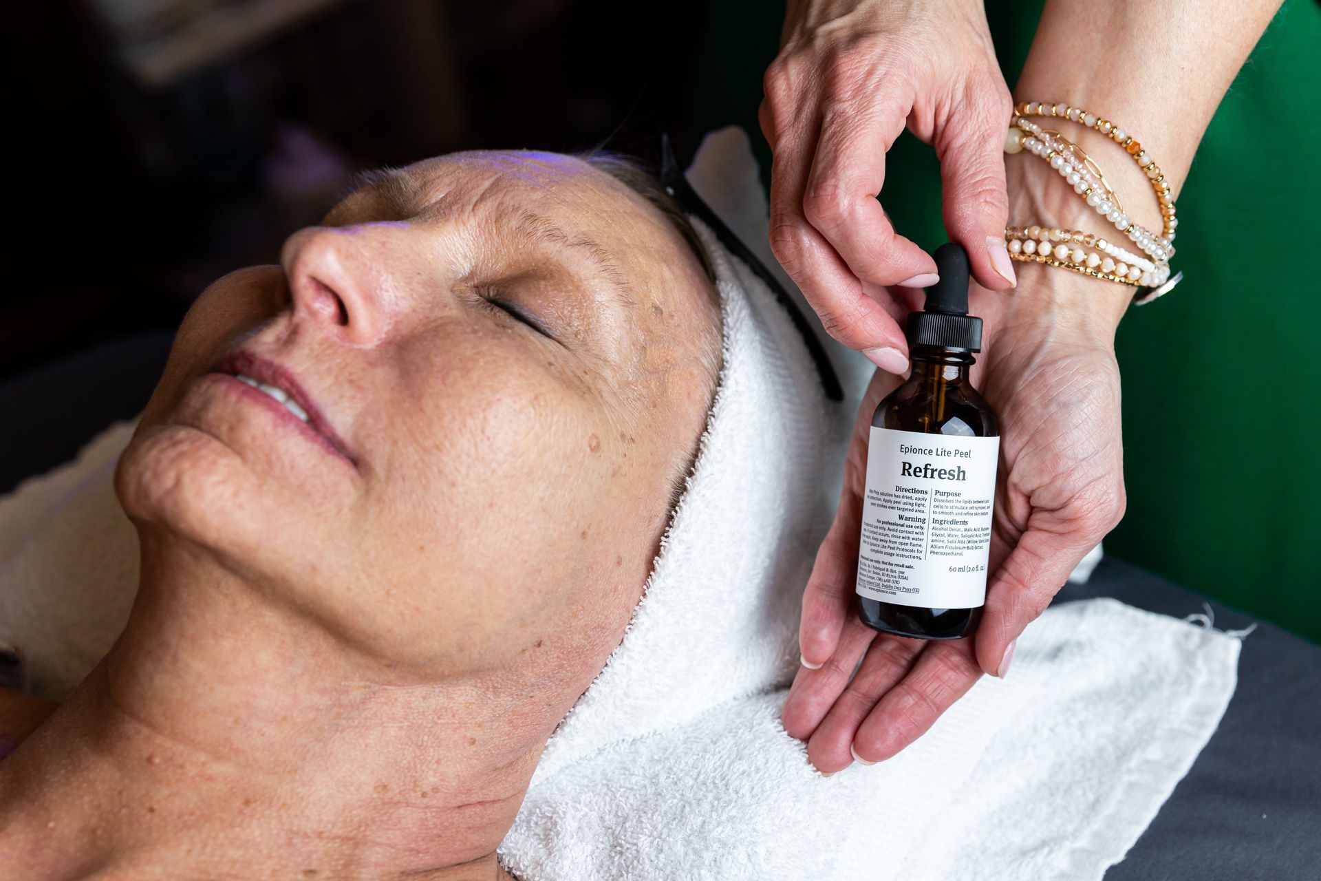 A professional applies a dropper of facial serum onto a client resting on a spa treatment table.