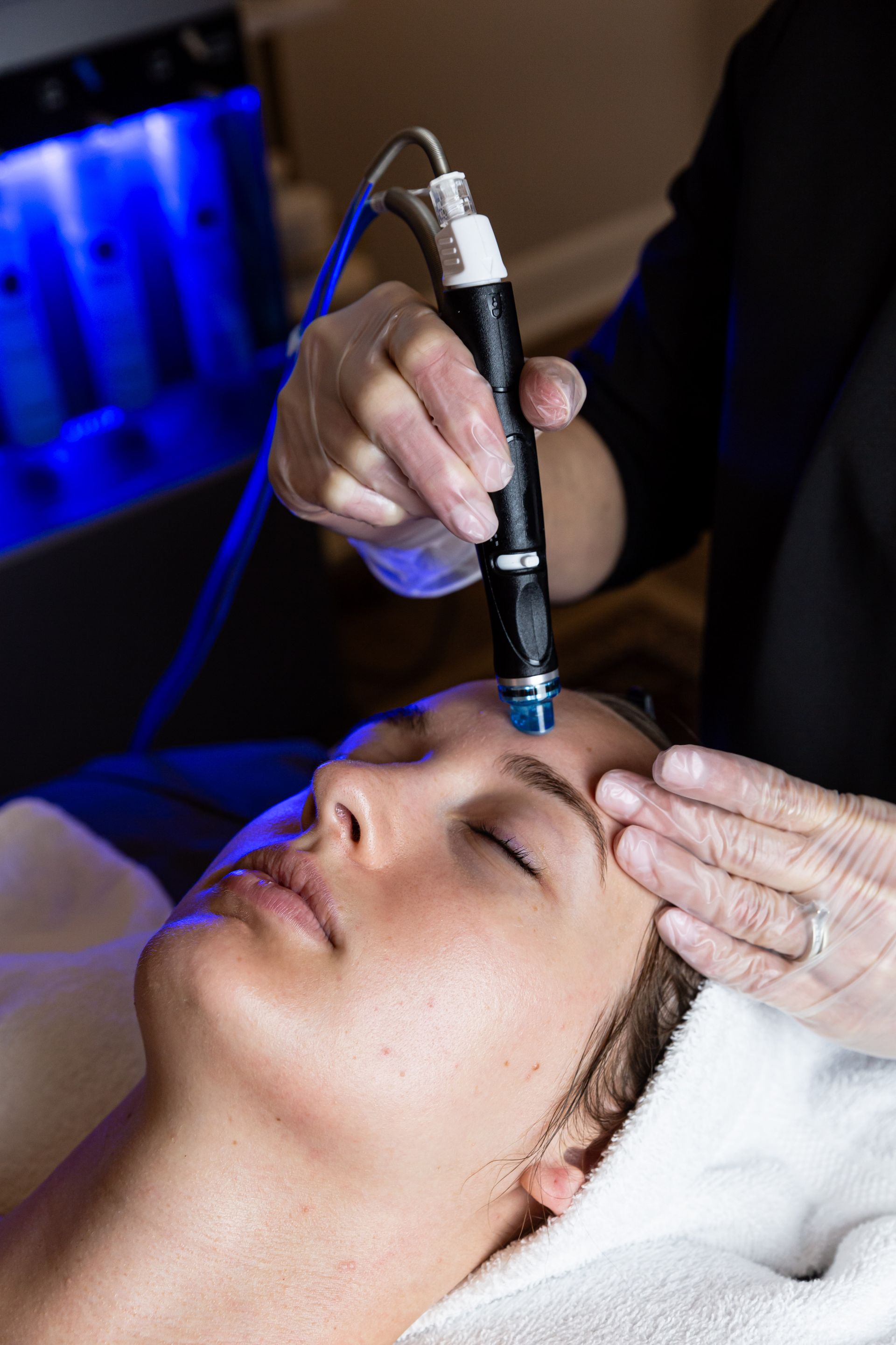 A professional uses a facial rejuvenation tool on a client’s forehead in a spa setting with blue ambient lighting.