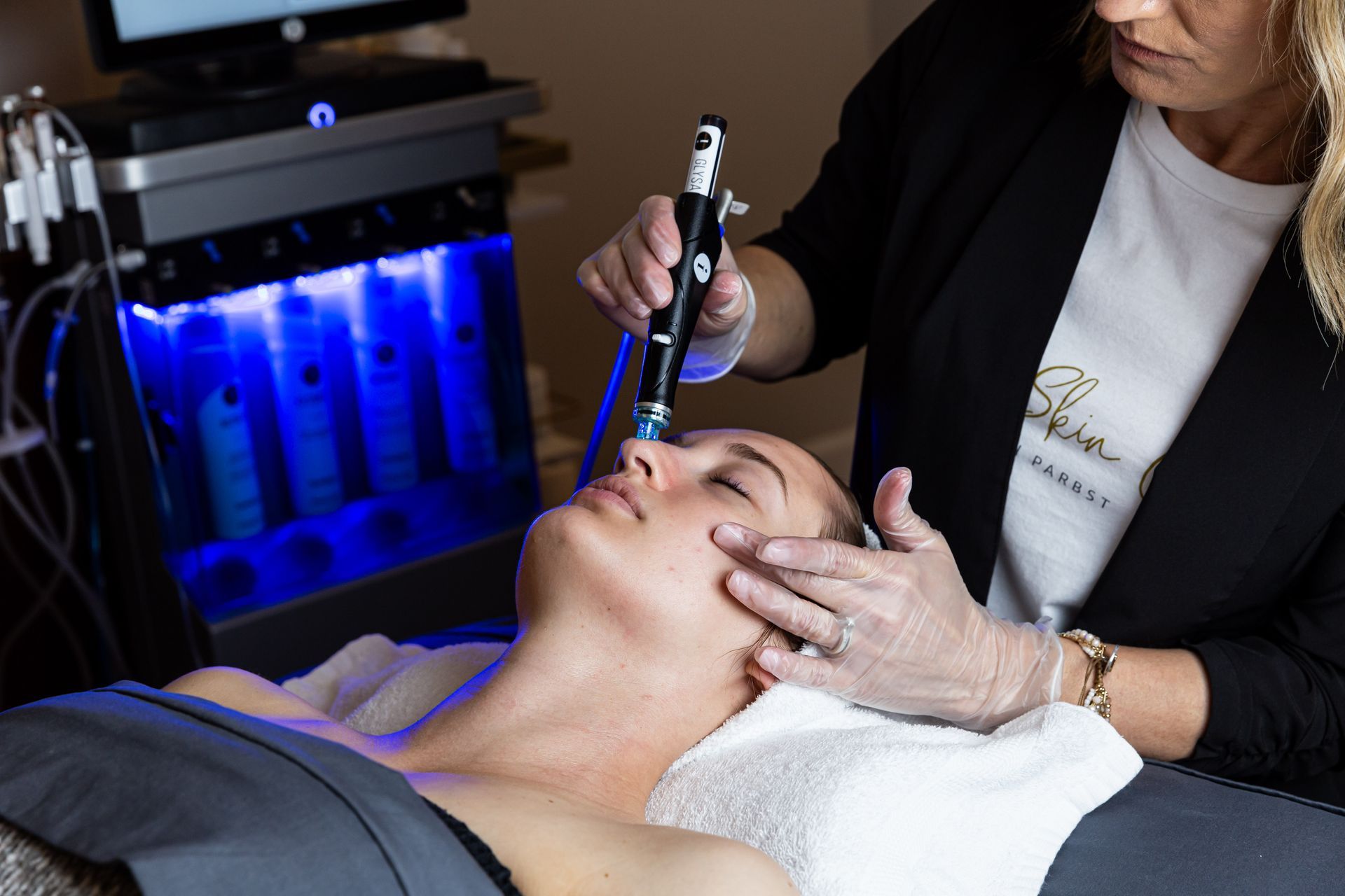 A professional performing a HydraFacial treatment on a client in a spa setting with a blue-lit machine in the background.