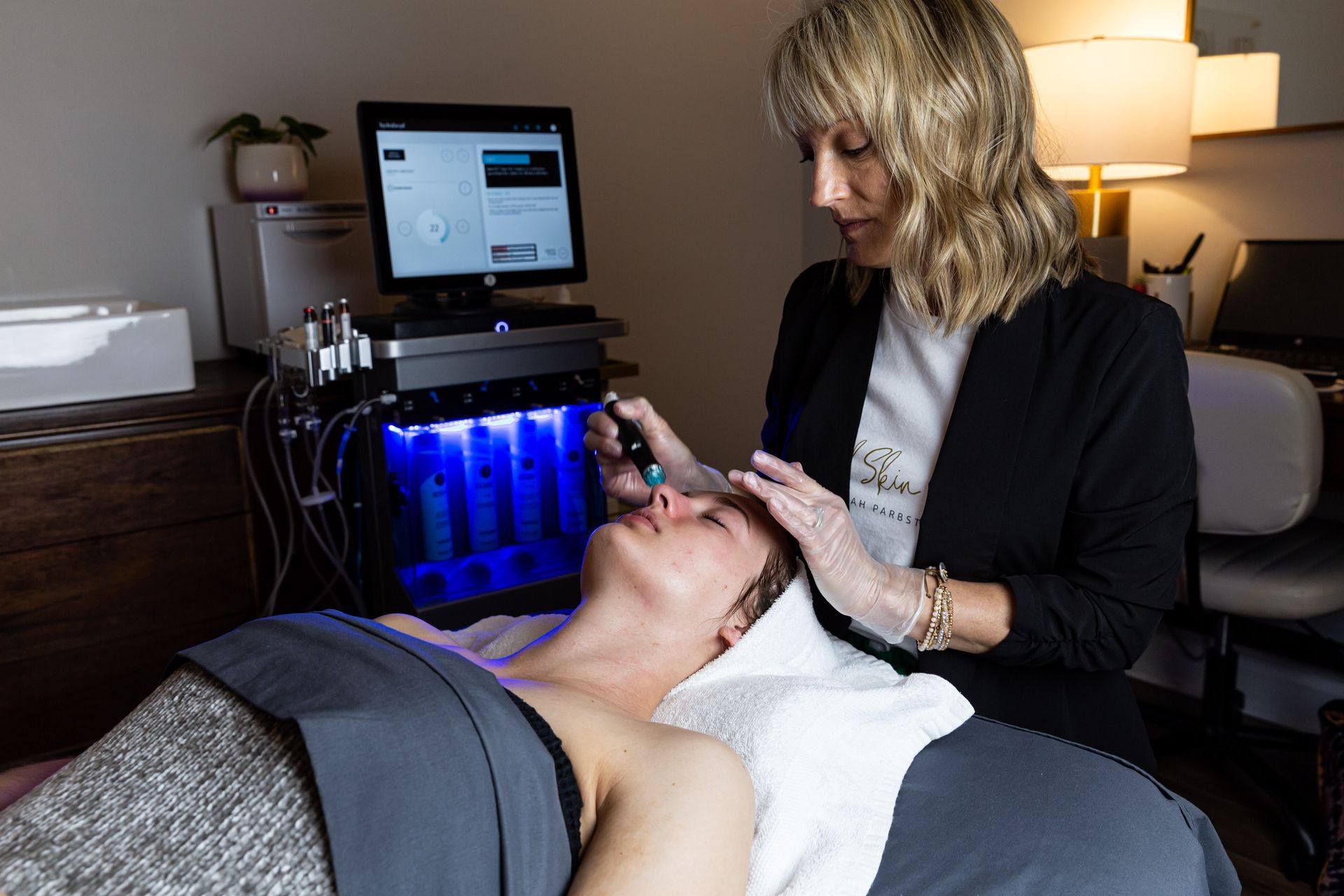 A professional performs a hydrafacial treatment on a client using a specialized machine with a glowing blue base.