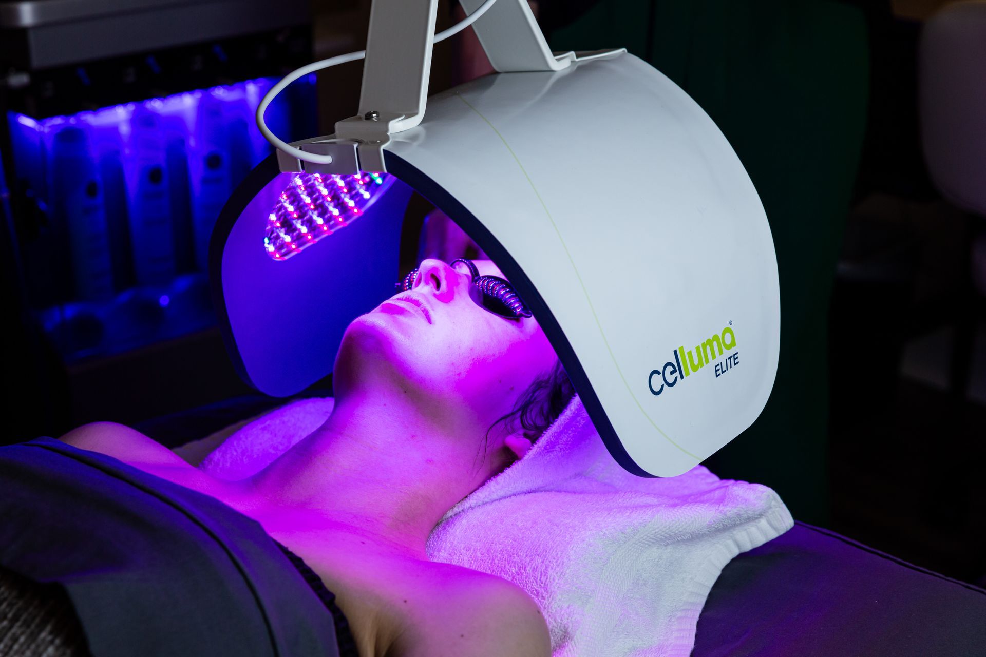 A person reclining under a curved LED light therapy device that is emitting a purple glow onto their face.