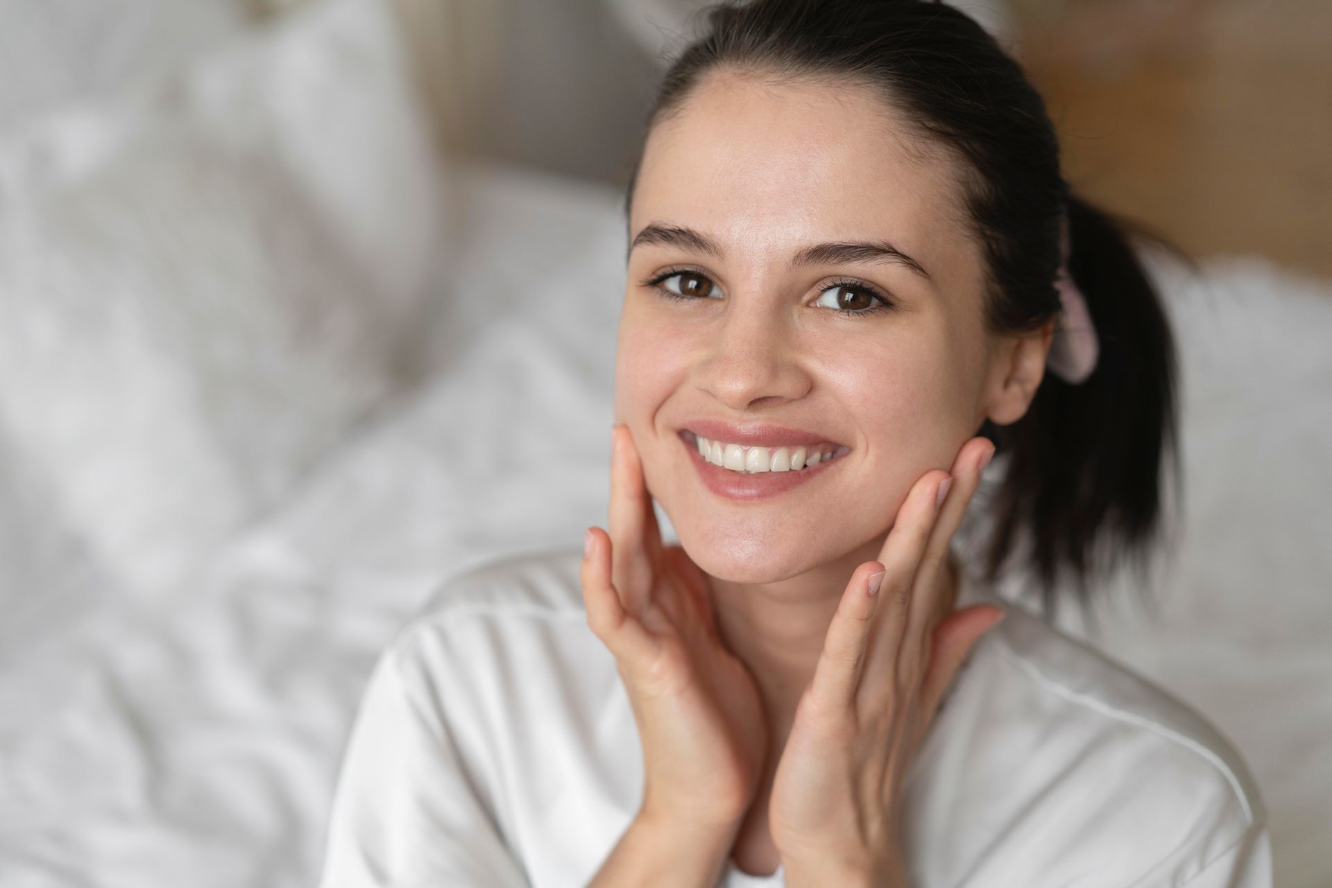 A person with a ponytail smiles while gently resting their hands on both sides of their face against a blurred background.