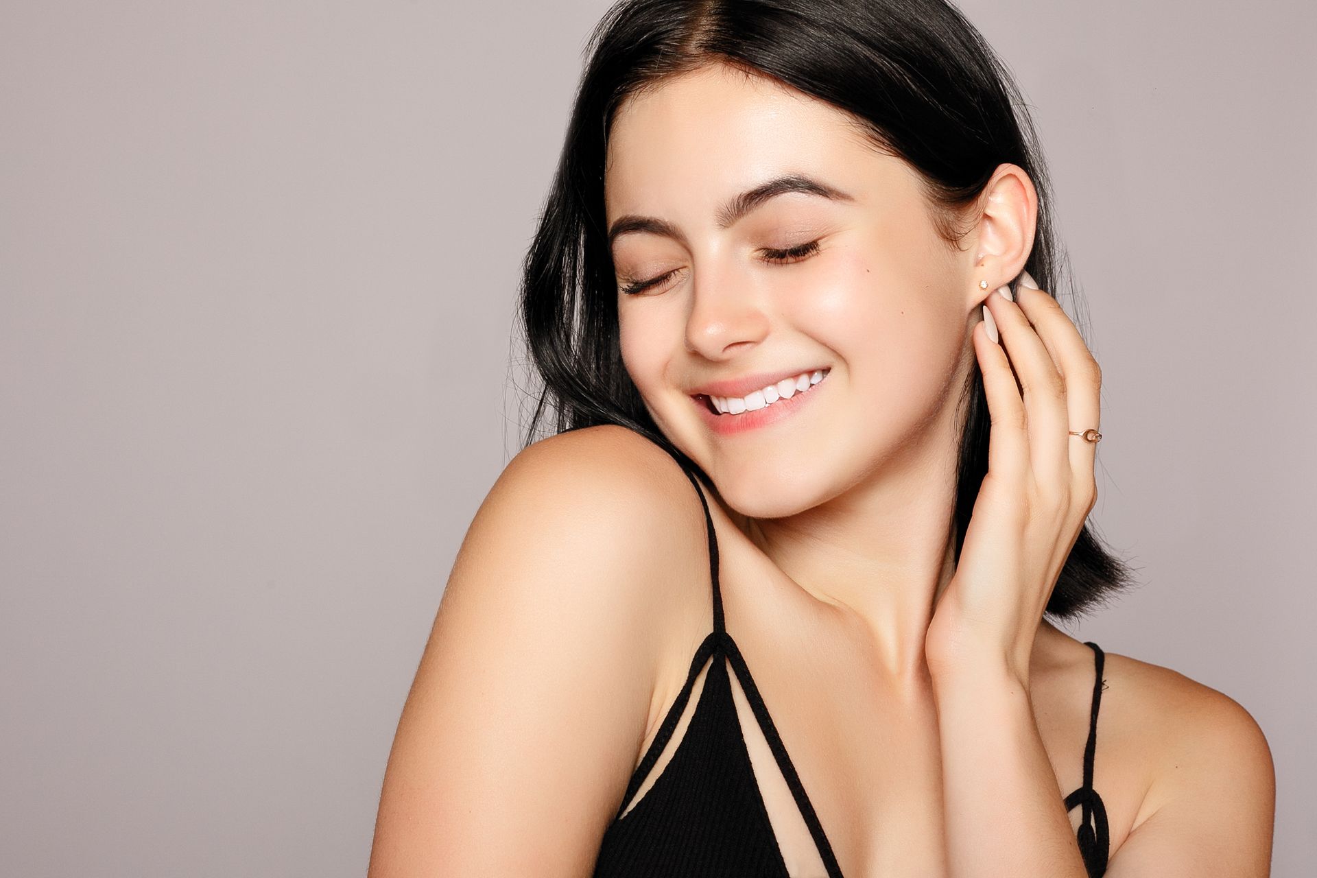 A person with dark hair smiling with eyes closed, touching their cheek against a plain light grey background.
