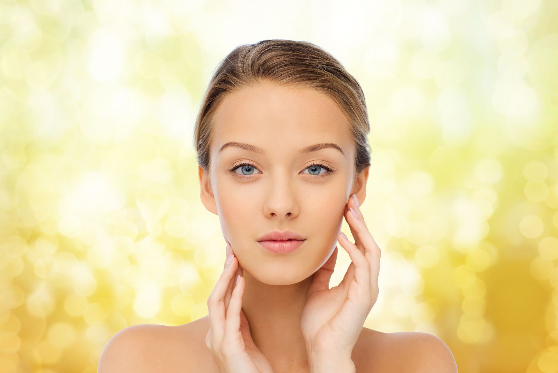 A person with clear skin touching their face with both hands against a soft-focus, golden, glowing background.