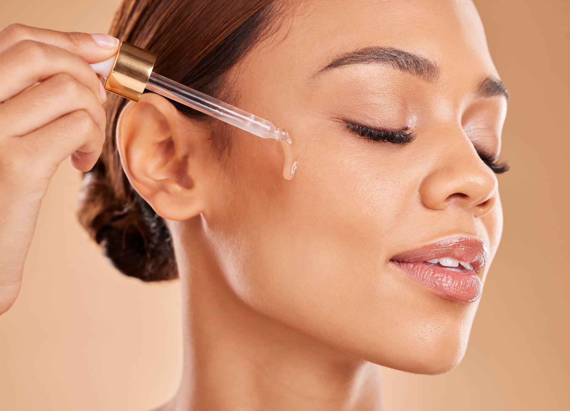 Close-up of a woman applying serum to her cheek with a dropper, eyes closed.