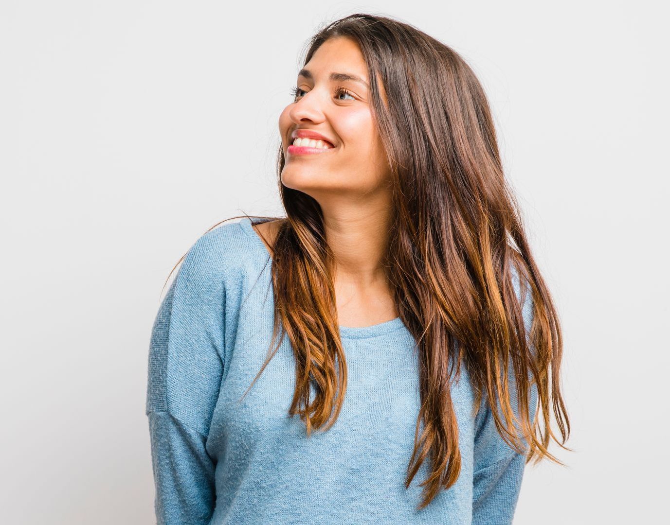 A person with long brown hair wearing a blue sweater smiles and looks toward the upper left against a white background.