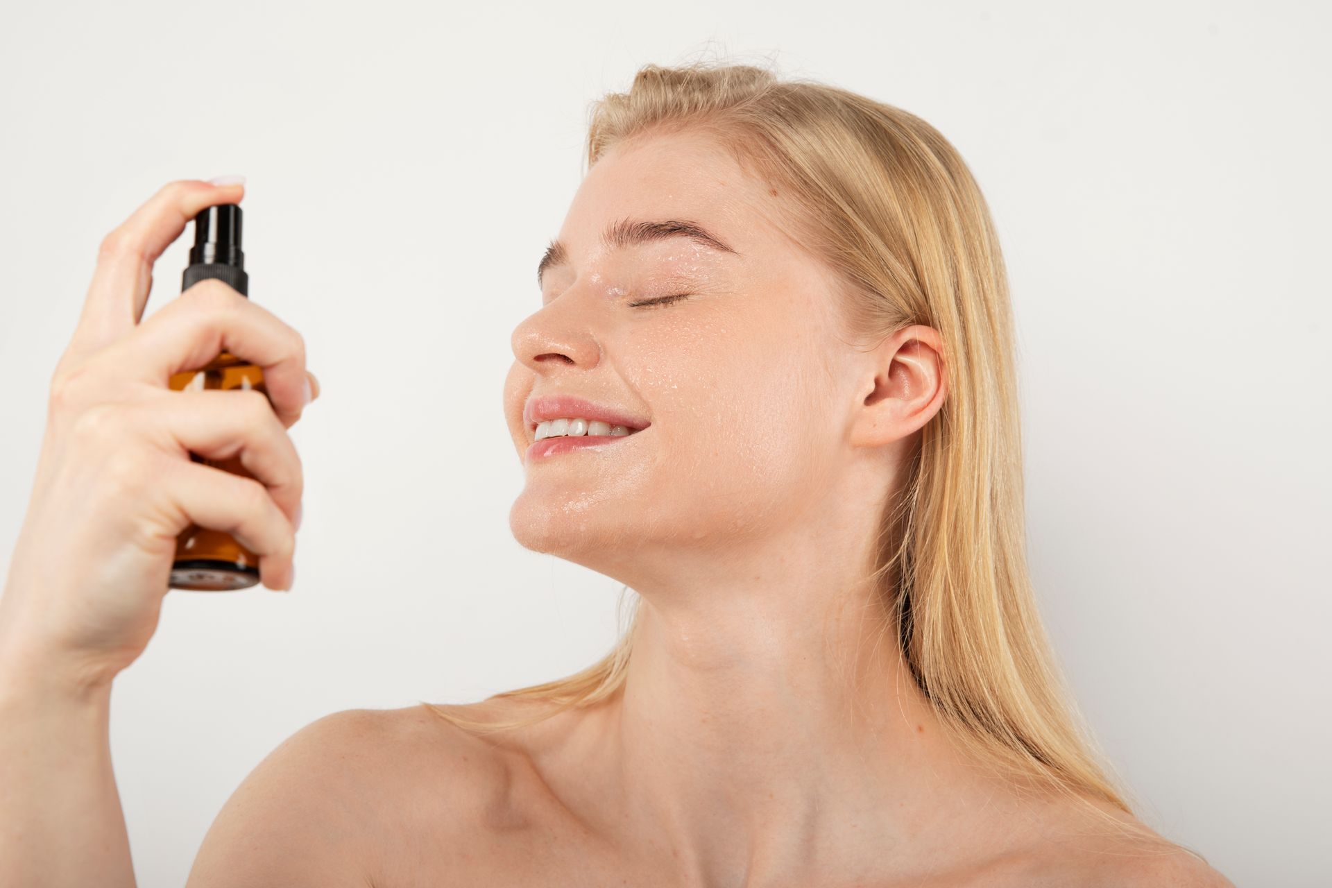 A person with their eyes closed smiling while spraying a mist from a small brown bottle onto their face.