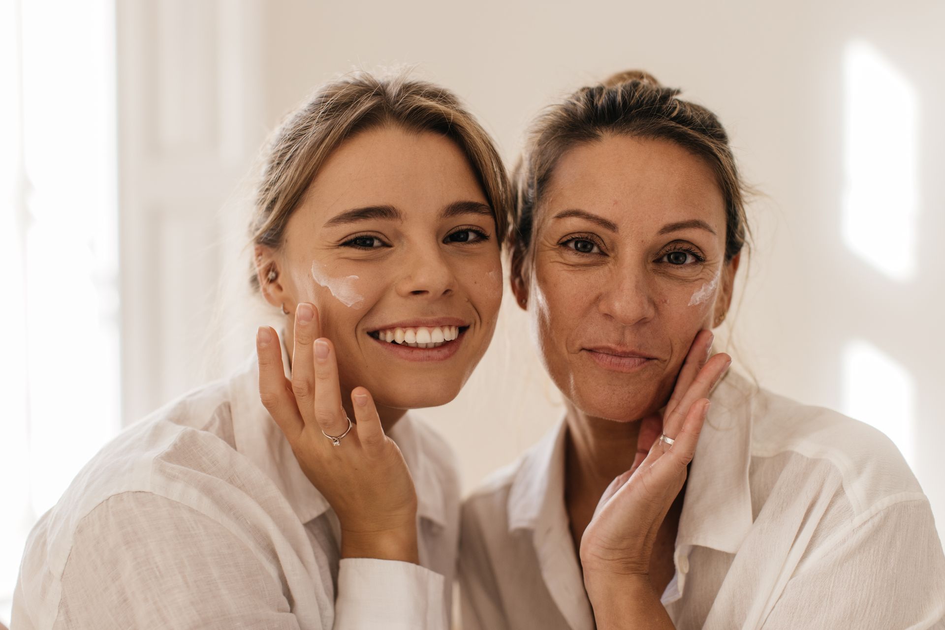 Two smiling individuals wearing white shirts applying skincare cream to their cheeks in a bright, indoor setting.