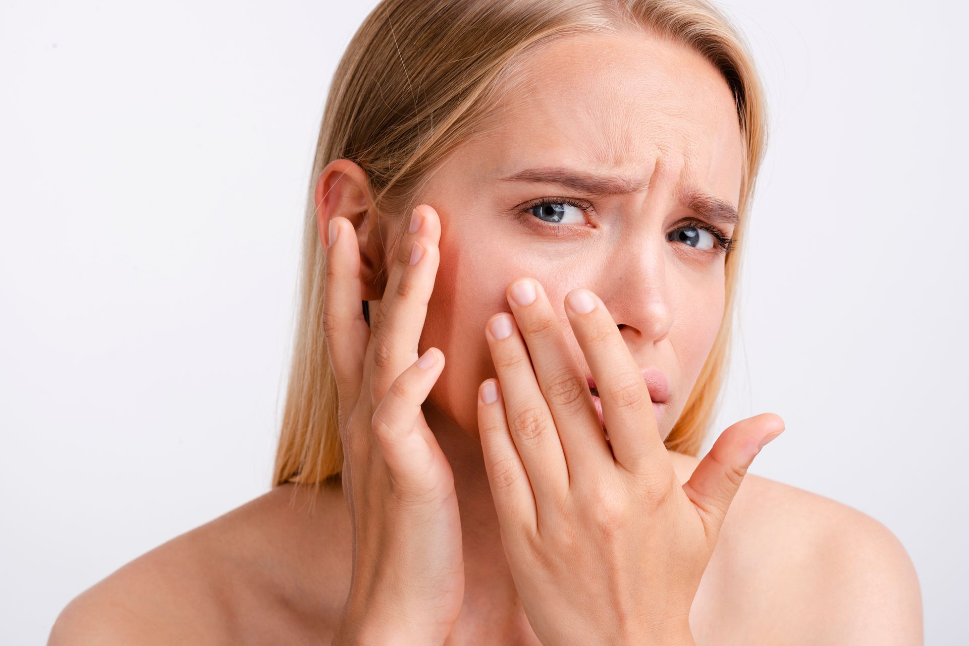 A person with a concerned expression gently touches their cheek, looking at their skin against a plain white background.