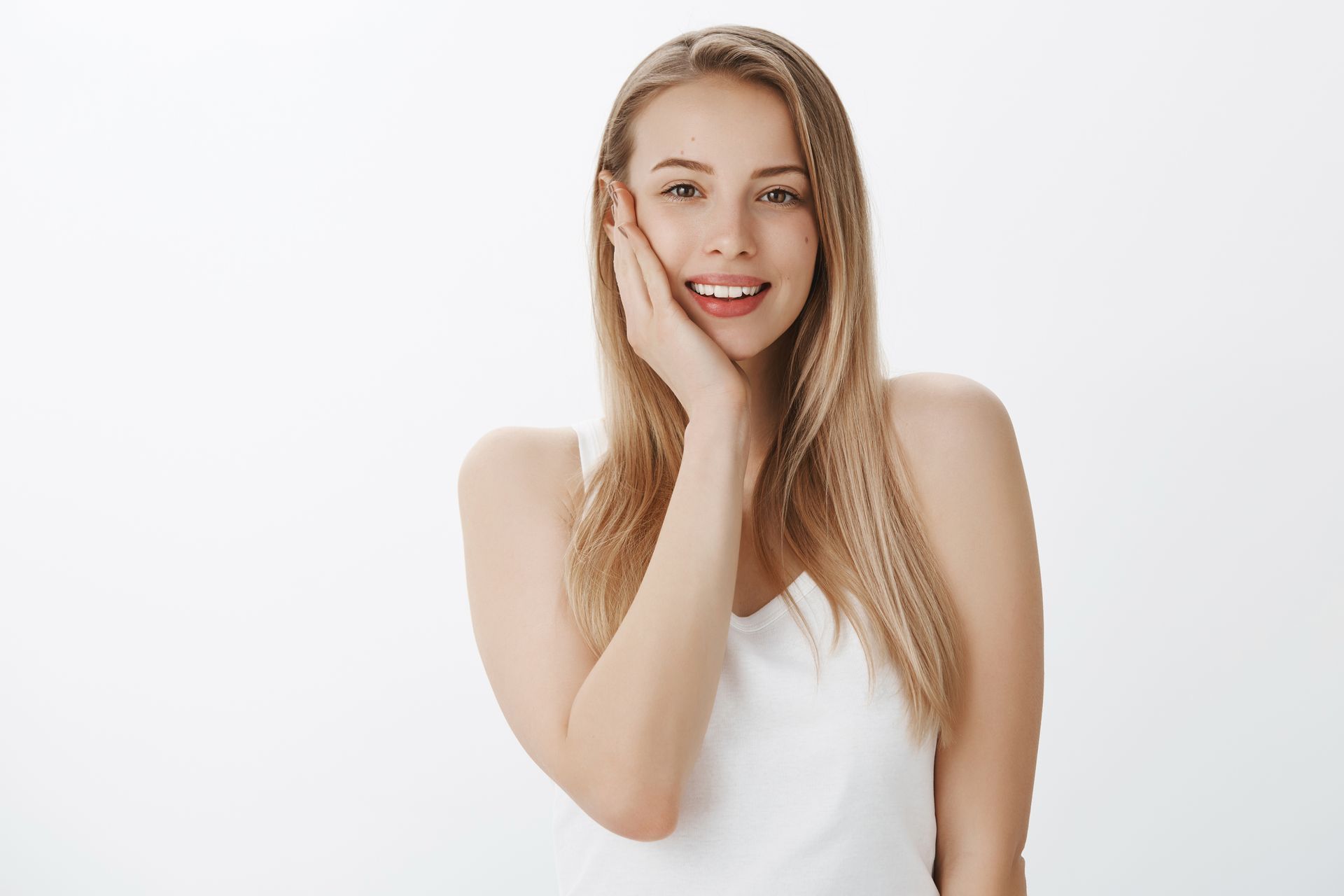 A smiling person with long blonde hair wearing a white tank top, resting their cheek on their hand against a white backdrop.