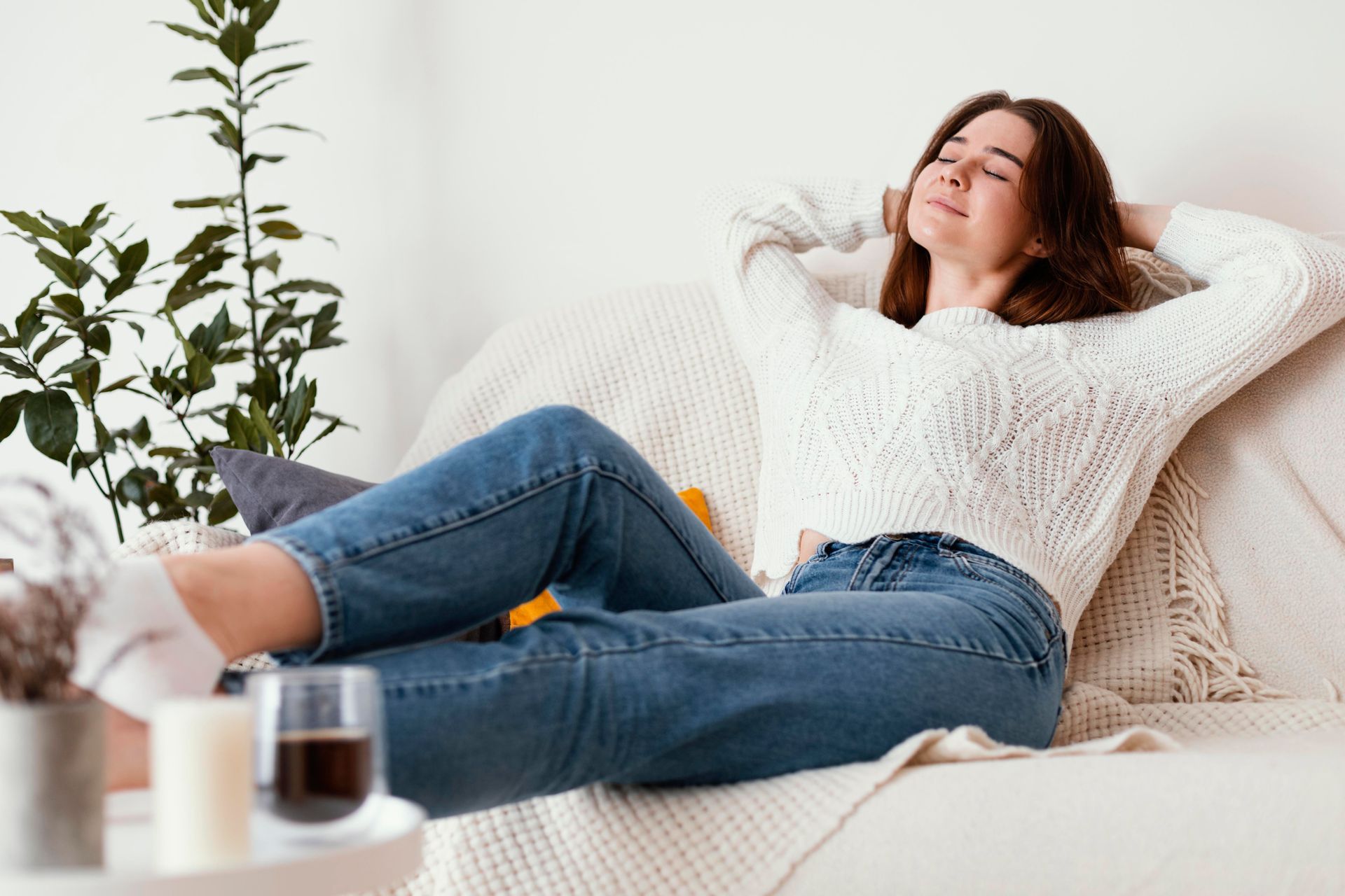 A person wearing a white sweater and blue jeans relaxes on a couch with eyes closed and hands behind their head.
