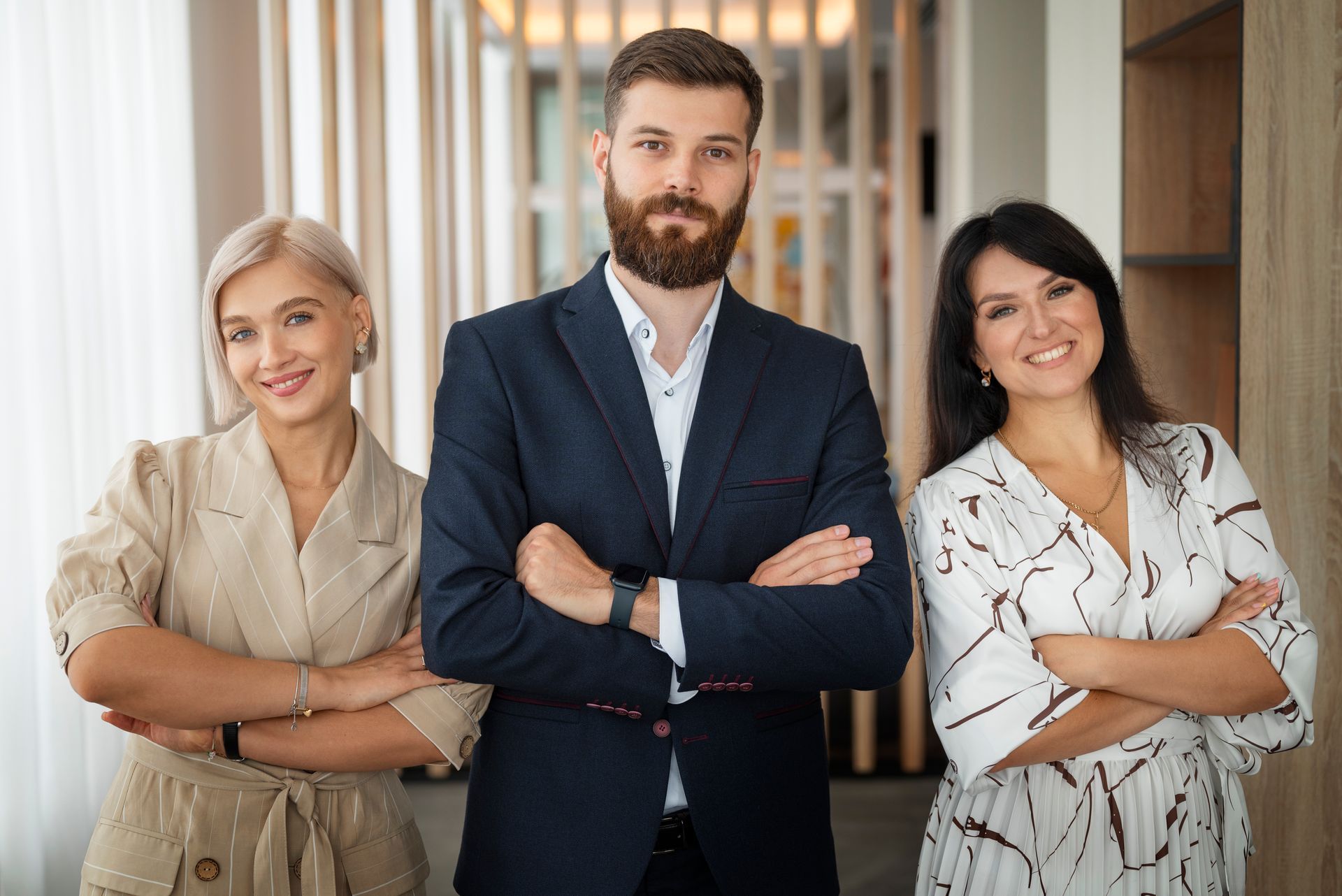 Three professionals stand side-by-side with arms crossed in an office hallway, smiling toward the camera.