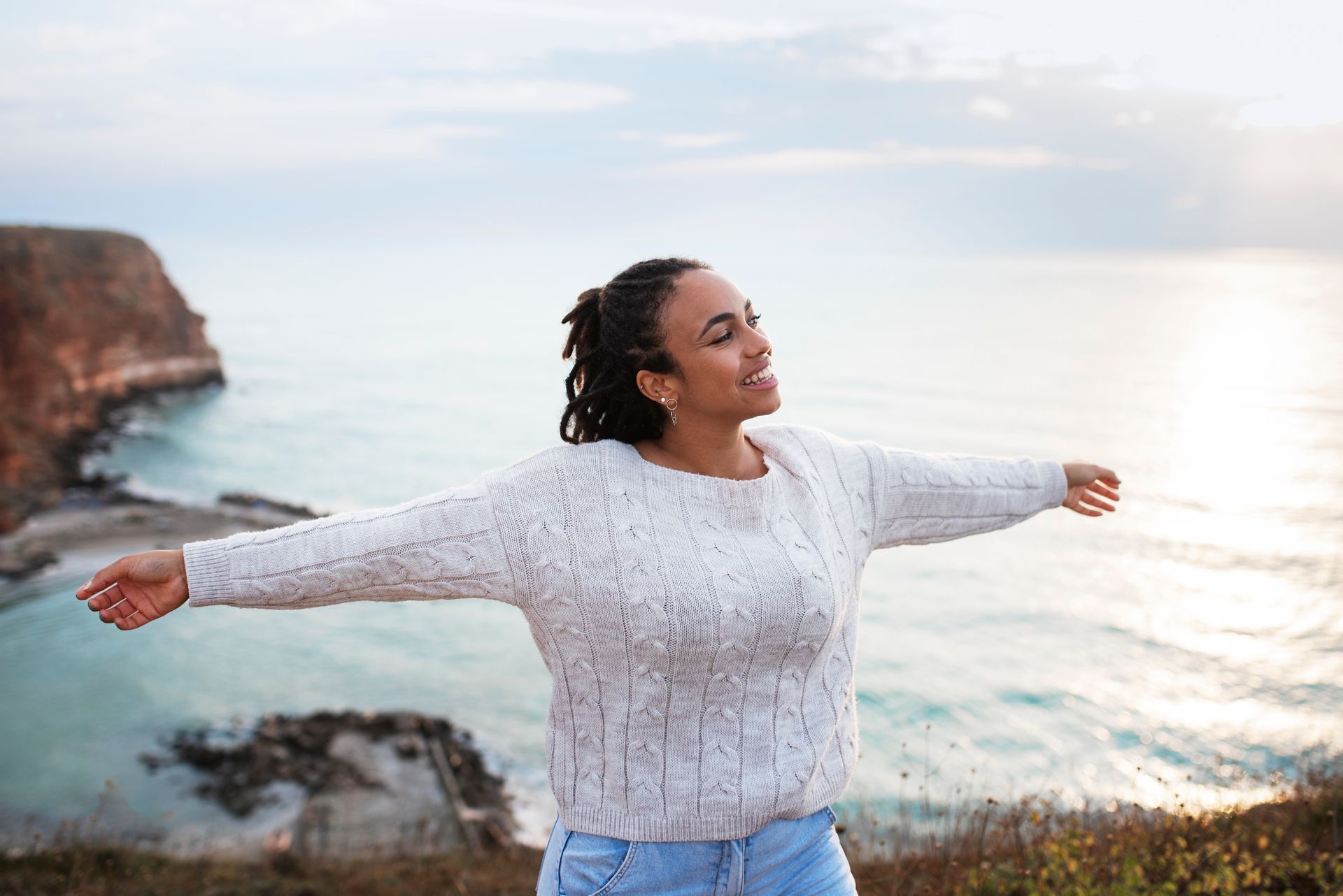 A person with braided hair stands on a cliff overlooking the ocean at sunset, arms wide, eyes closed with a calm expression.