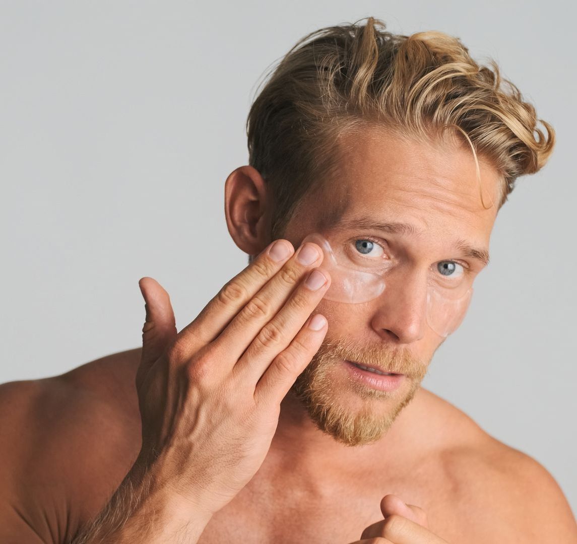 A person with blonde hair applies an under-eye mask to their cheek against a neutral gray background.