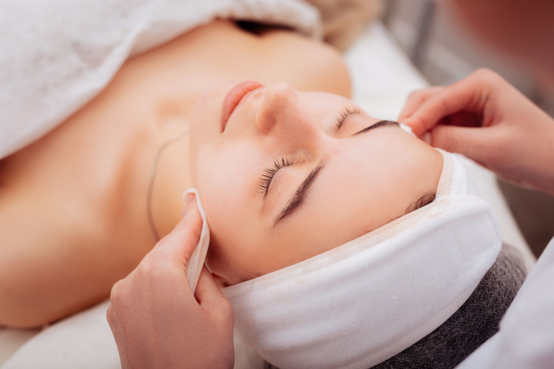 A professional aesthetician cleanses a client's face with a cotton pad while the client relaxes with eyes closed.