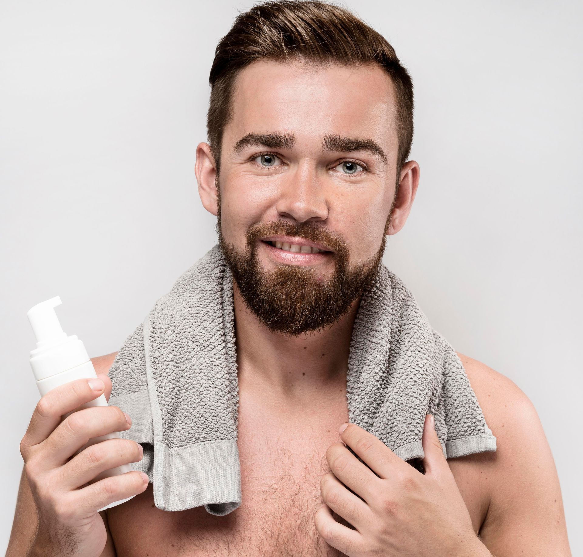 A person with a beard and a towel around their neck smiling while holding a white pump bottle against a neutral background.