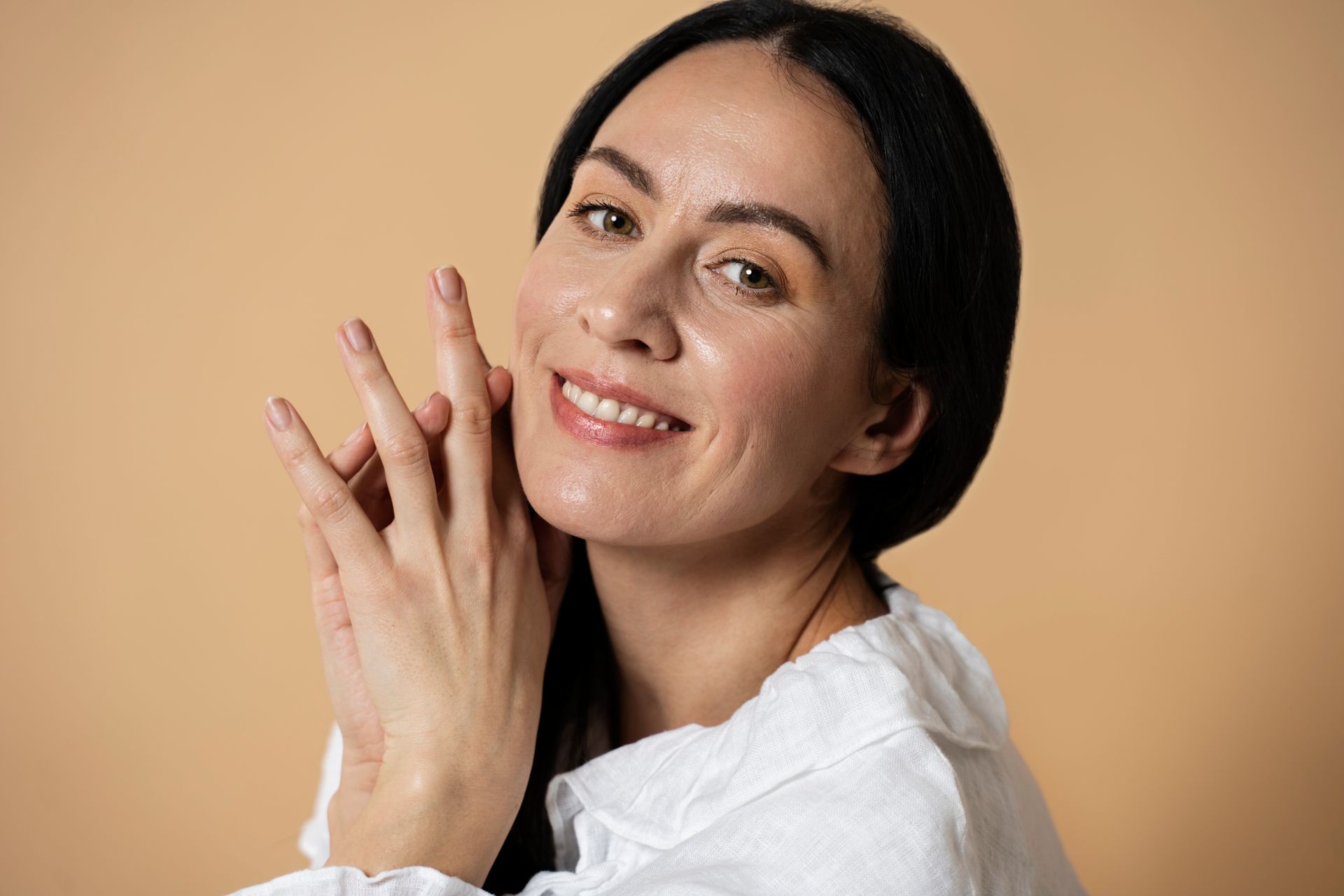A smiling person with dark hair wears a white shirt, posing with hands near their cheek against a beige background.