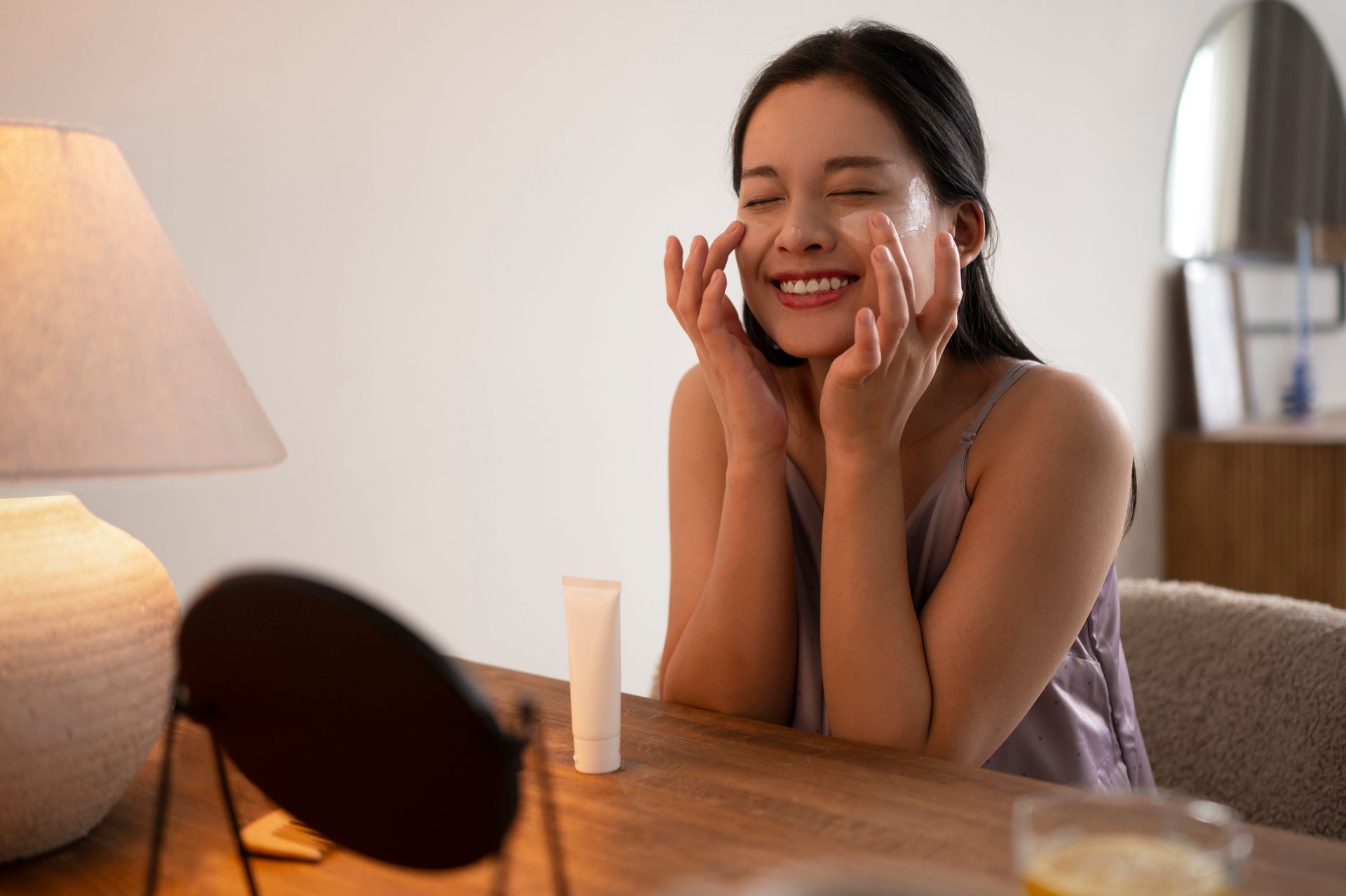 A person sits at a table, smiling while applying cream to their cheeks in front of a mirror and a lamp.