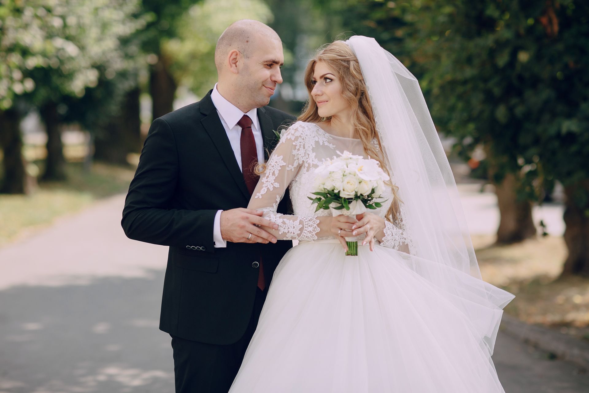 A bride in a white lace dress holds a bouquet while looking at the groom, who wears a black suit with a dark red tie.