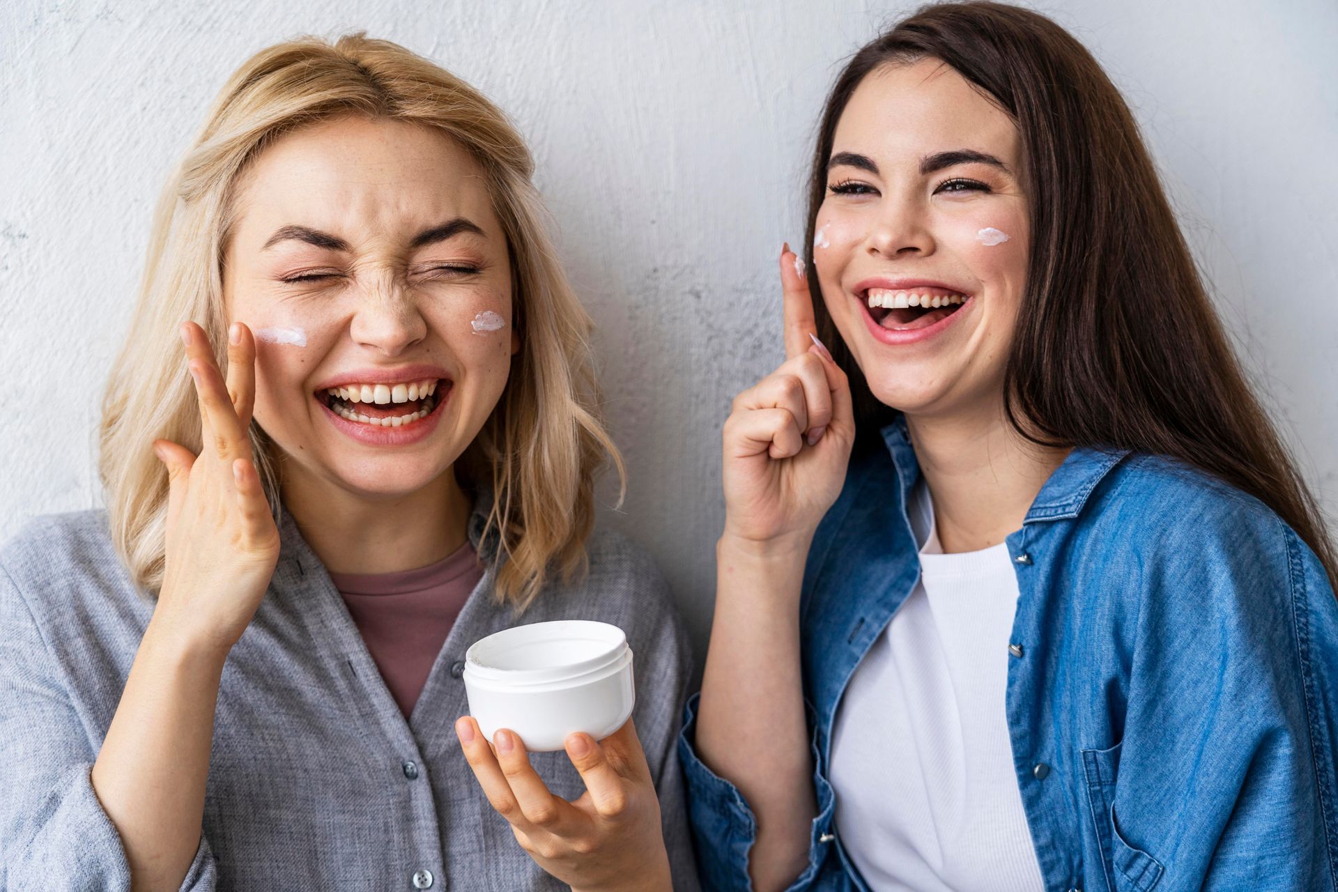 Two smiling people applying white cream to their cheeks against a white background.