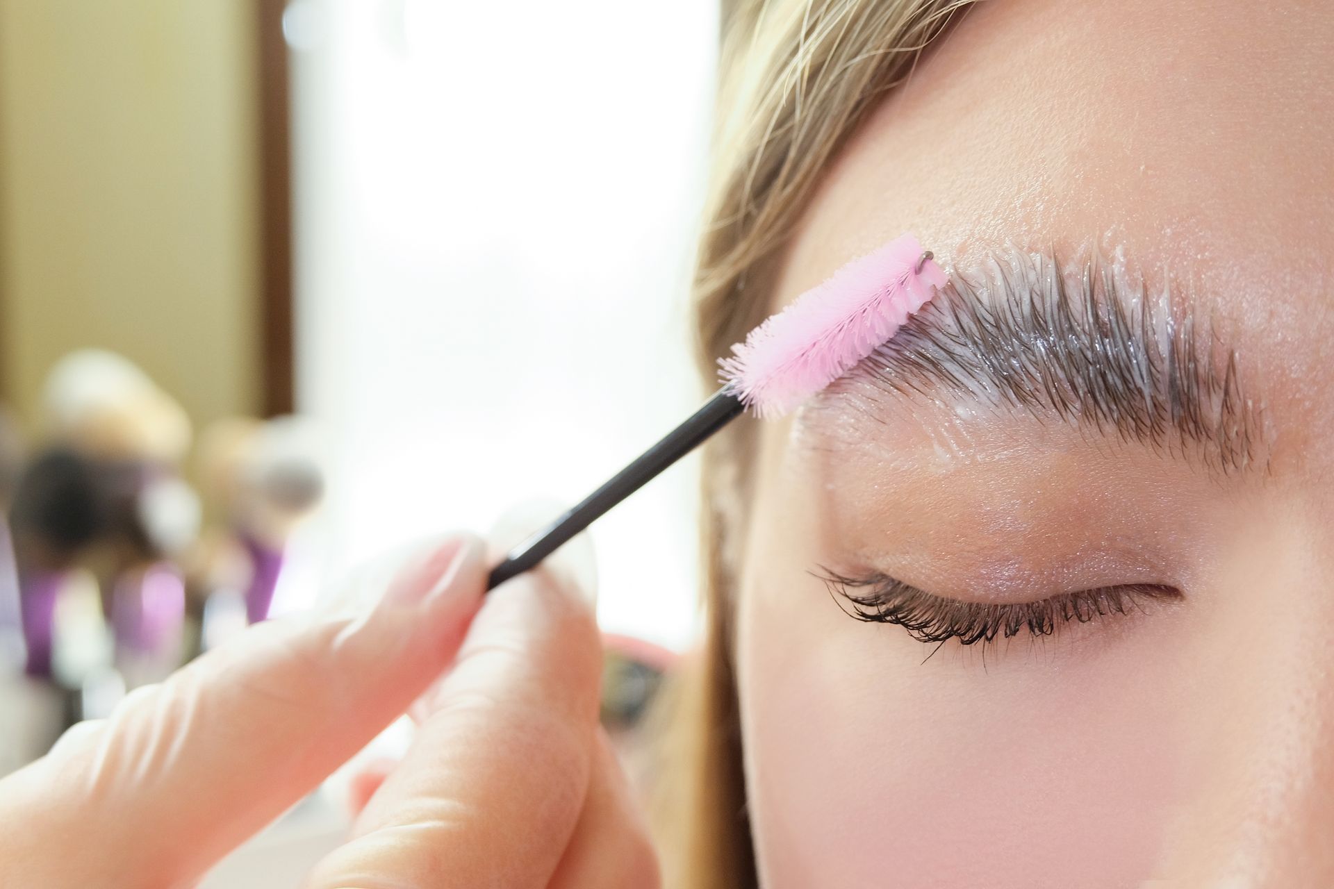 A person receives an eyebrow lamination treatment, with a technician using a pink spoolie to brush product onto the brow.