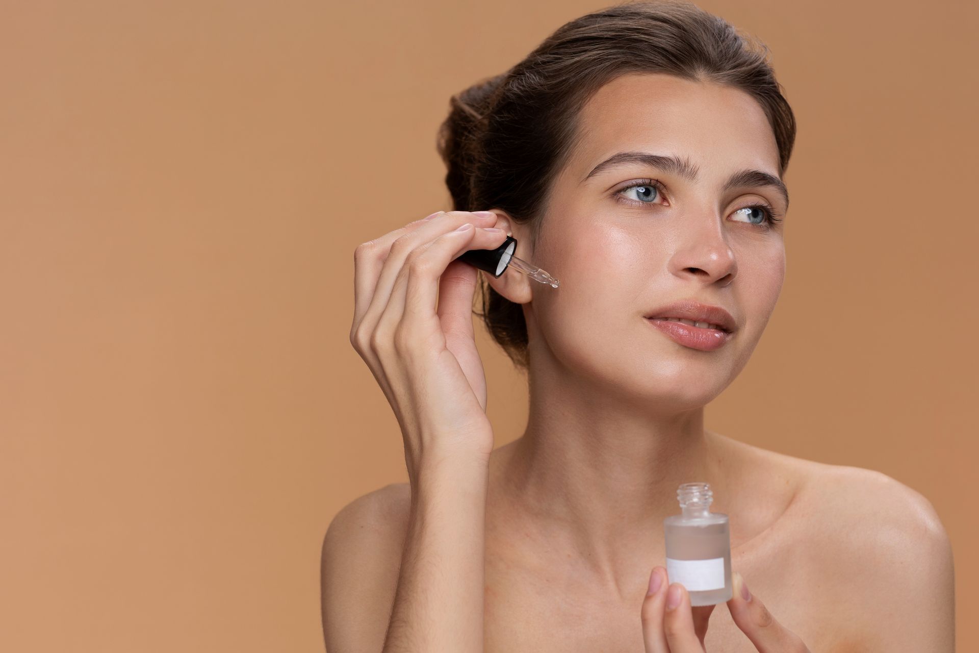 A person applying serum from a glass dropper to their cheek against a tan background.