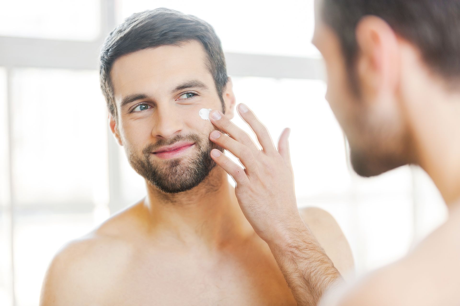 A person looks in a mirror and applies a small dot of white cream to their cheek.