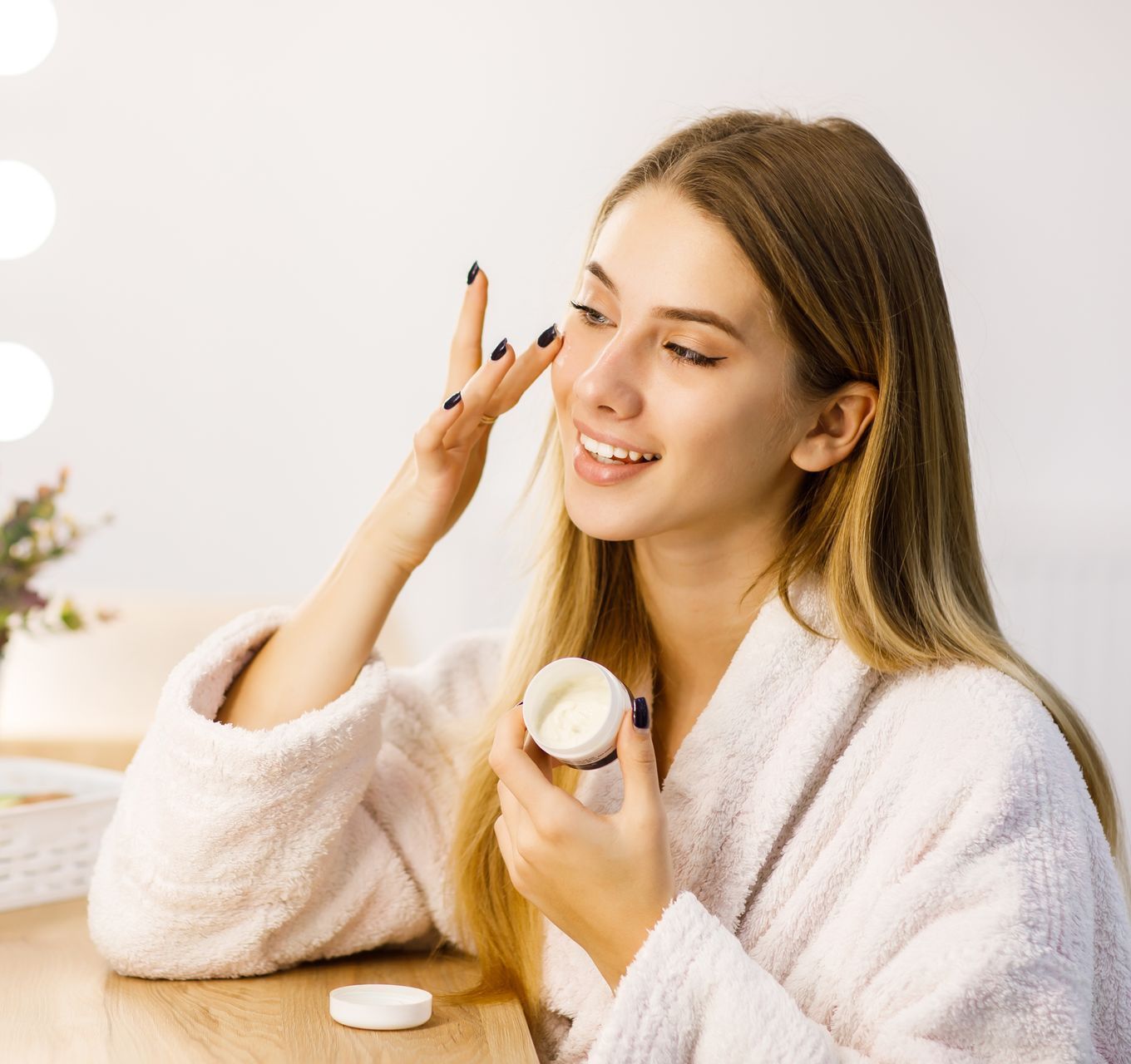 A smiling person in a white robe applies facial cream from a small jar, sitting at a vanity table with vanity mirror lights.