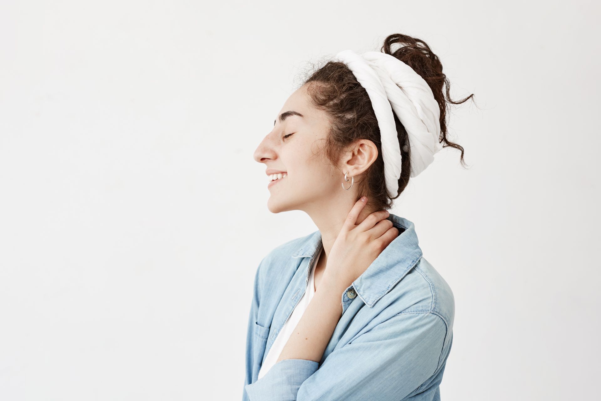A person with curly hair wrapped in a white towel smiles with eyes closed, touching their neck against a white background.